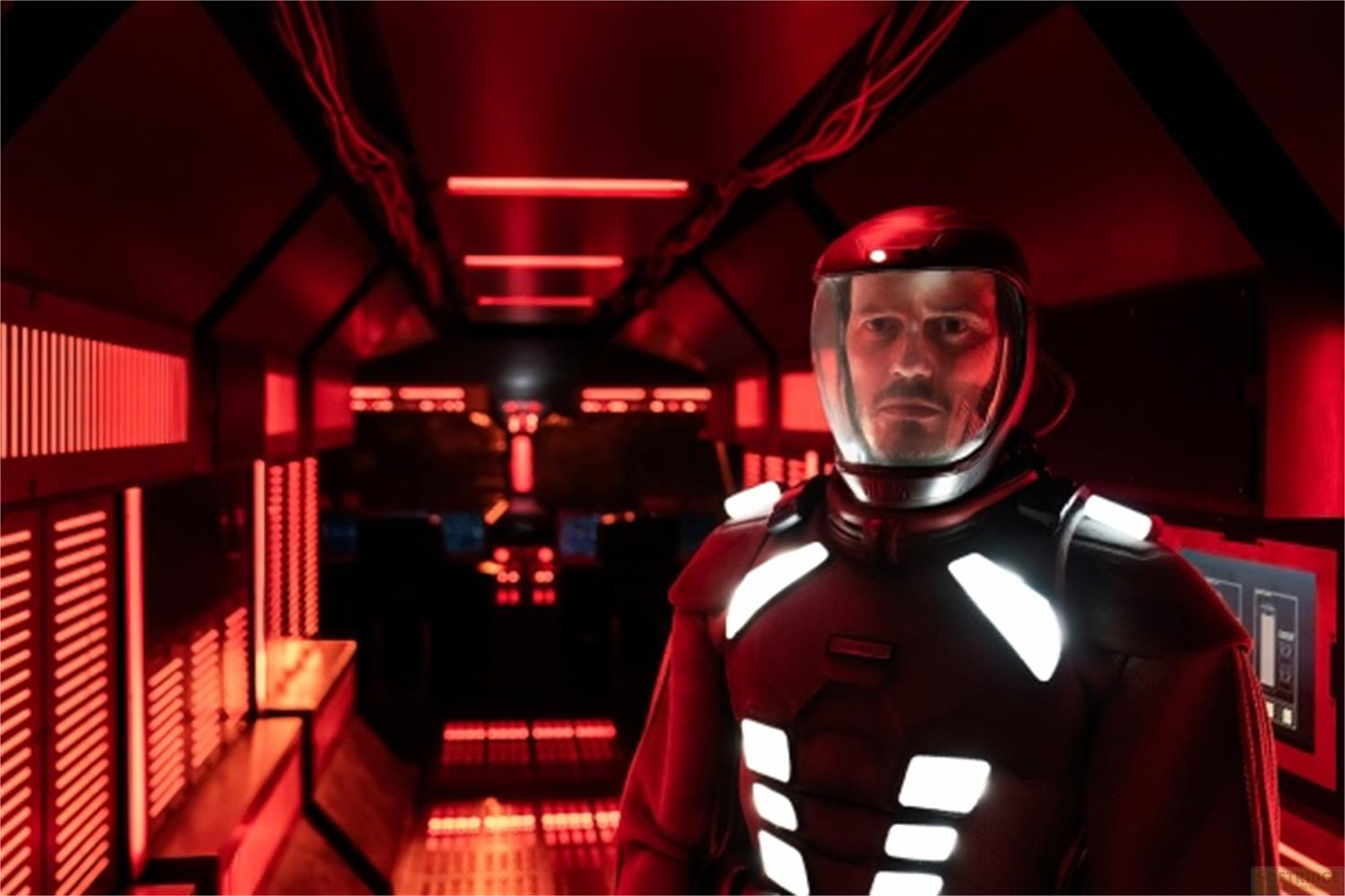 Un uomo in tuta spaziale scura con casco si trova in un corridoio rosso illuminato.