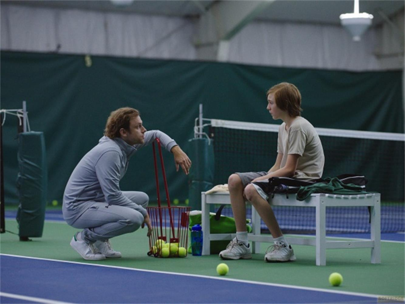Ein Trainer kniet neben einem Korb mit Tennisbällen und spricht mit einem Jungen.