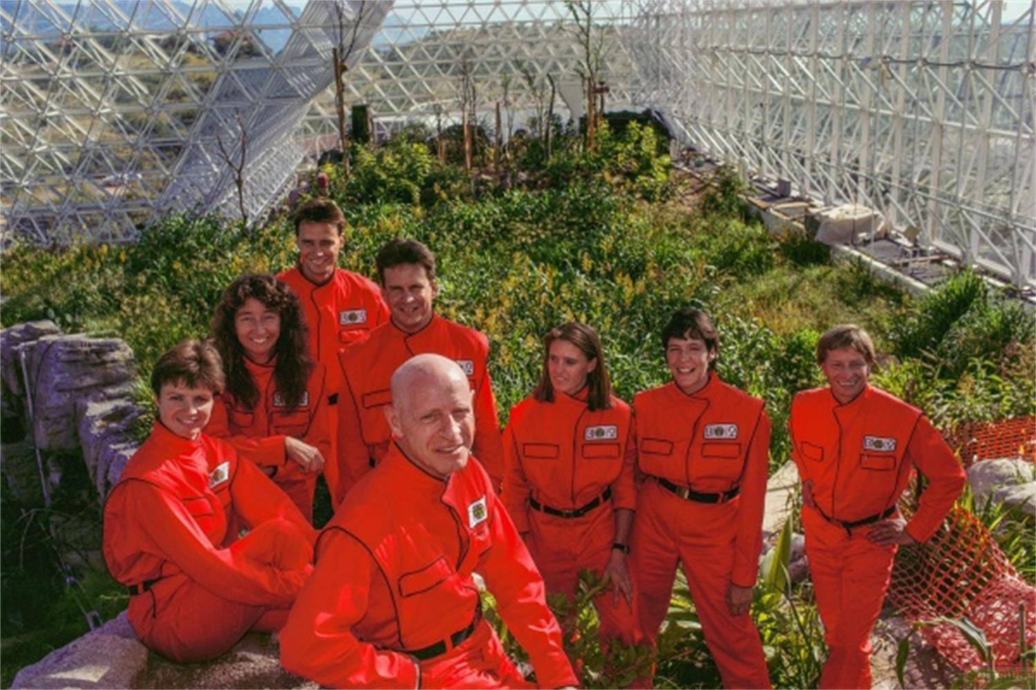 Otto persone in tute rosse si trovano in un ambiente verde lussureggiante sotto una cupola geodetica.