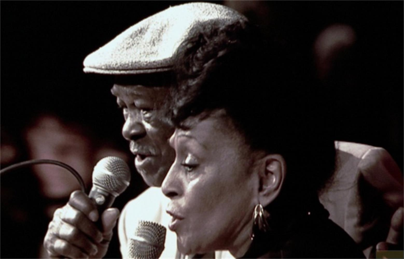 Un uomo con berretto e una donna cantano nel microfono.