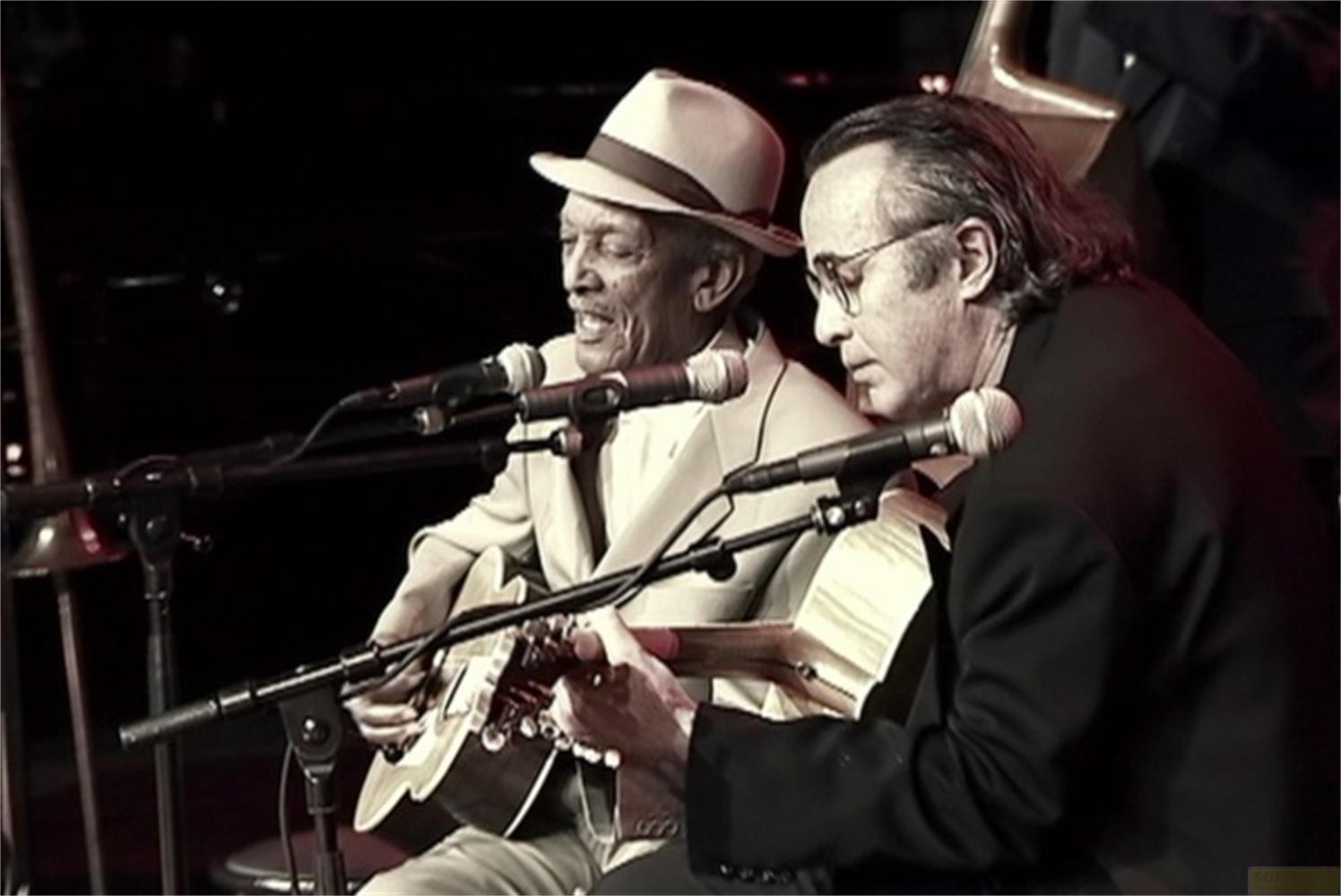 Due uomini suonano la chitarra e cantano nei microfoni; uno porta un cappello.