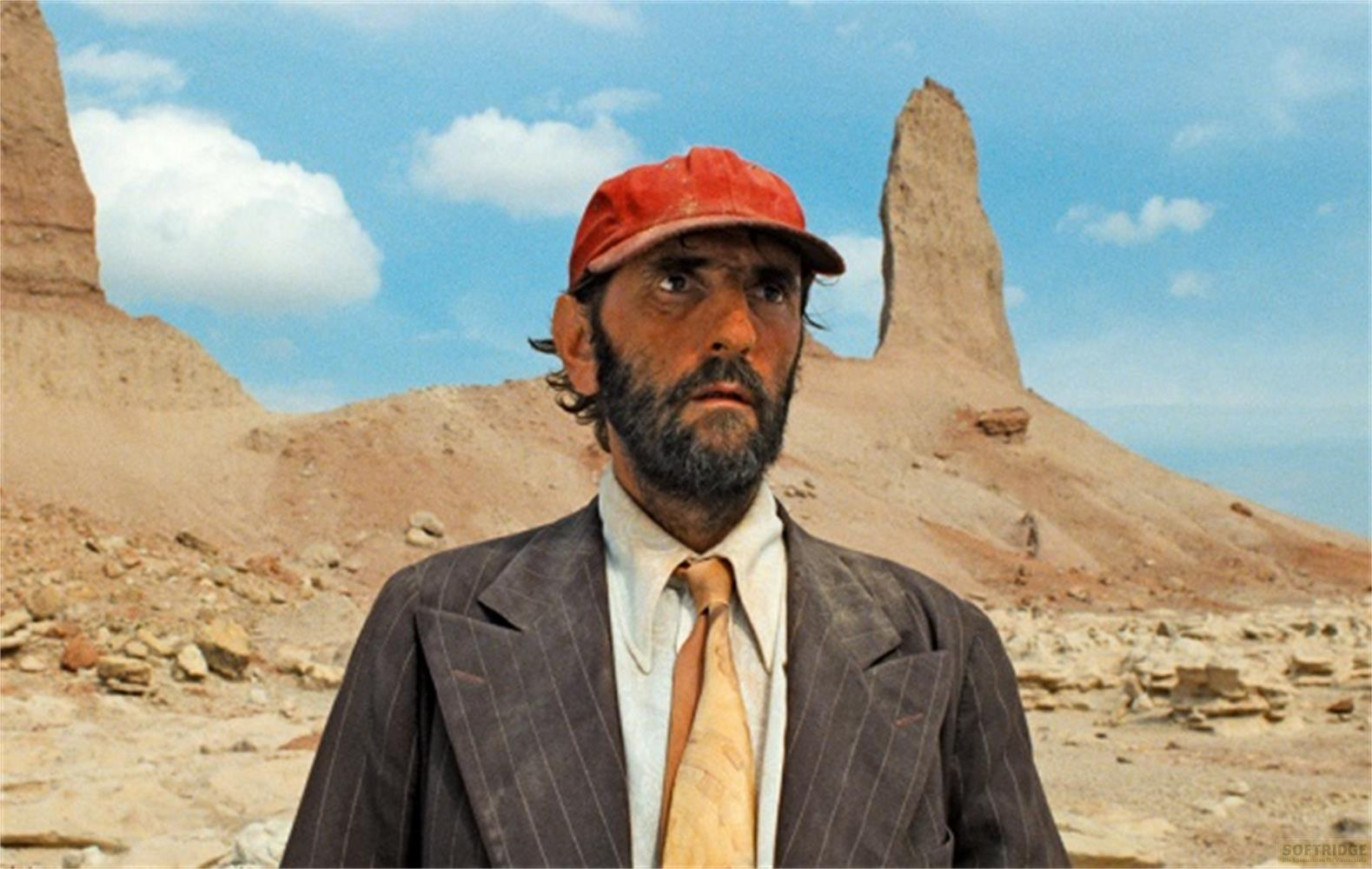 Un uomo barbuto, cappellino rosso, completo, paesaggio desertico.
