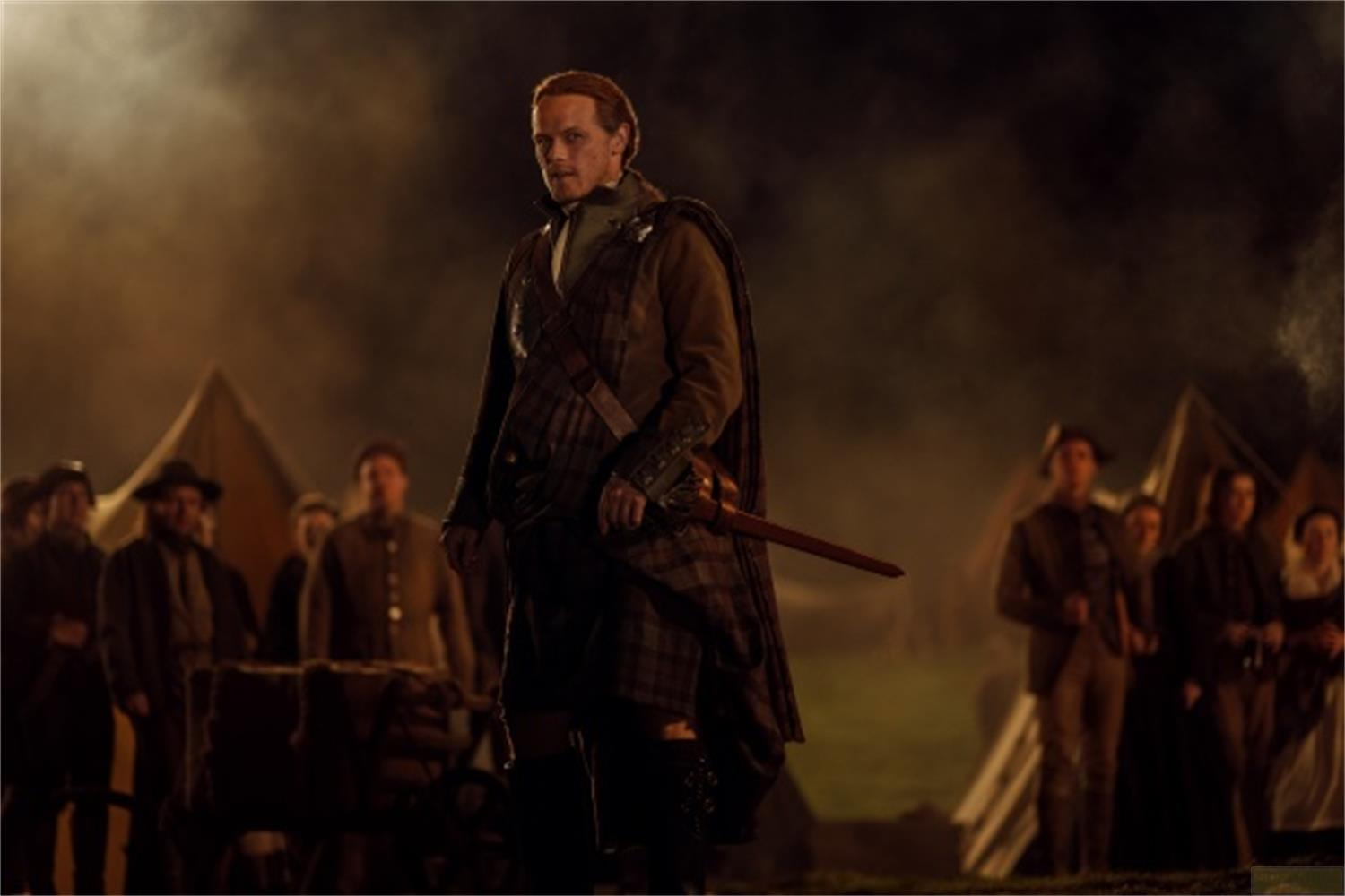 Un uomo in kilt e giacca marrone in primo piano. Tende e persone sullo sfondo.