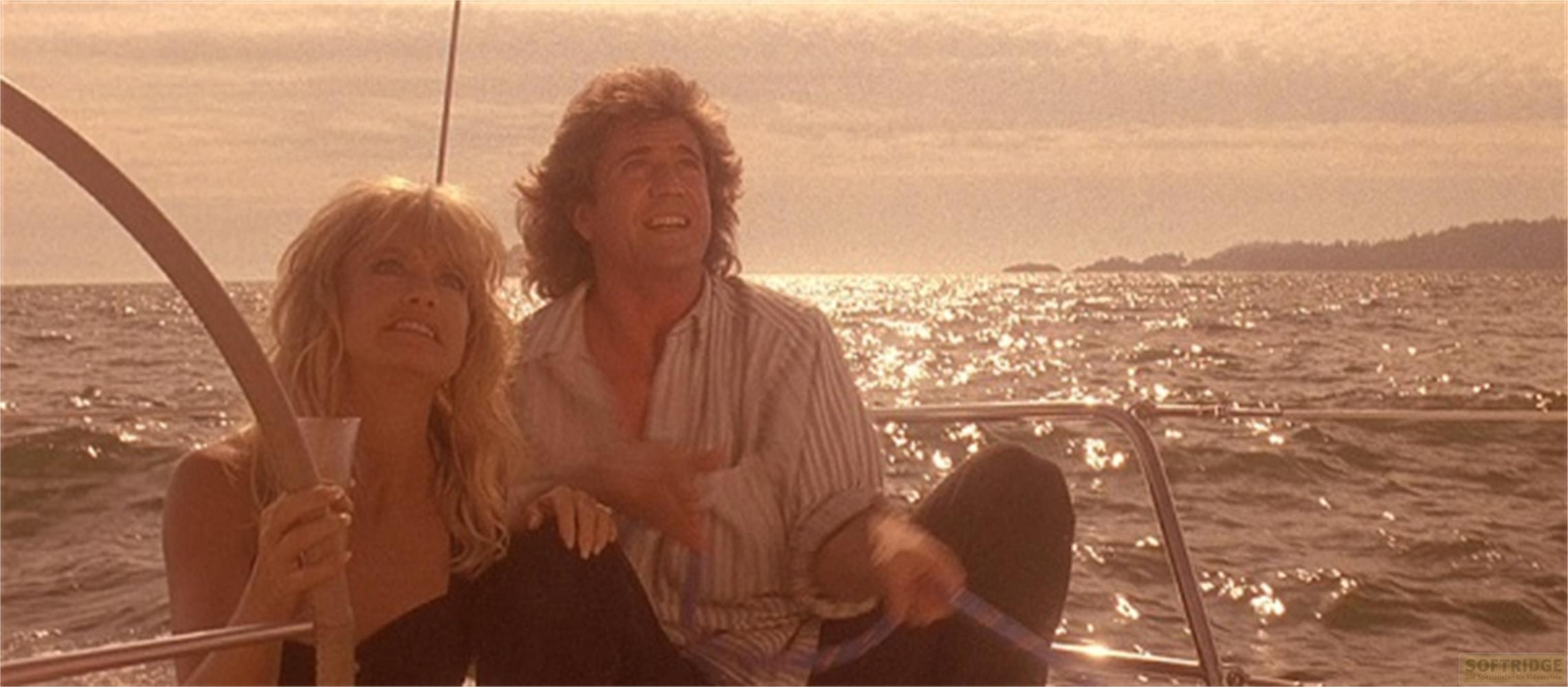 Un uomo e una donna su una barca a vela al tramonto, con una costa sullo sfondo.
