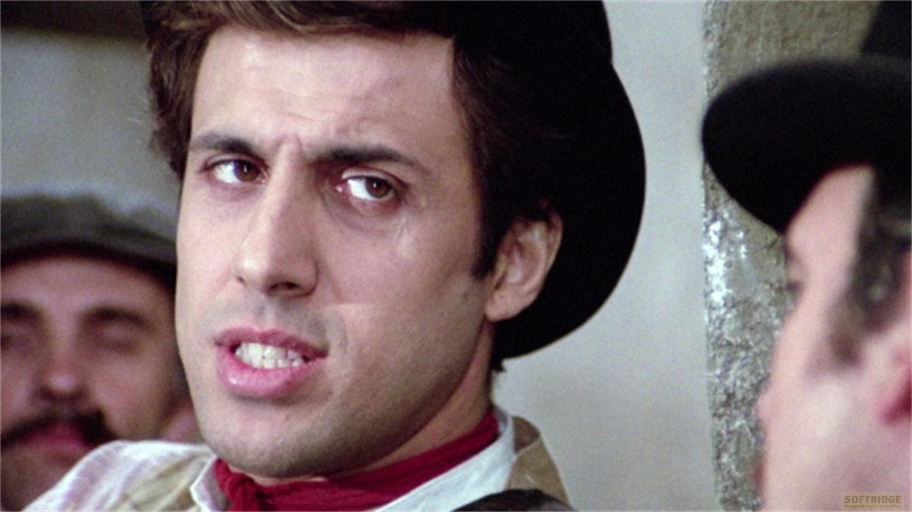 Uomo con cappello e sciarpa rossa guarda a sinistra. Un altro uomo con baffi sullo sfondo.