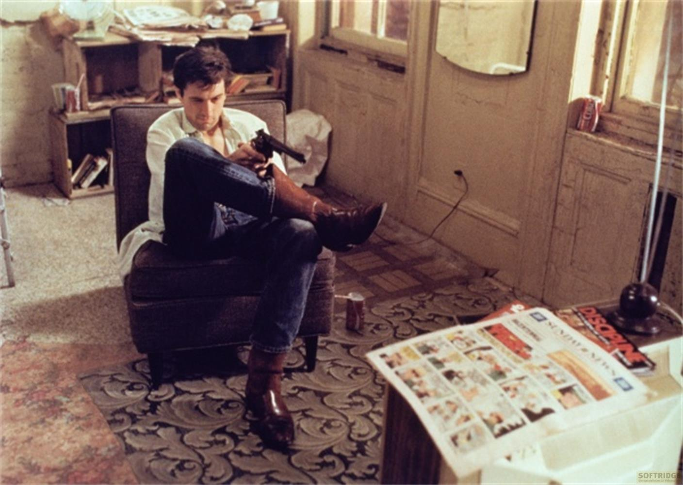 Homme assis dans un fauteuil avec un pistolet, journaux devant.