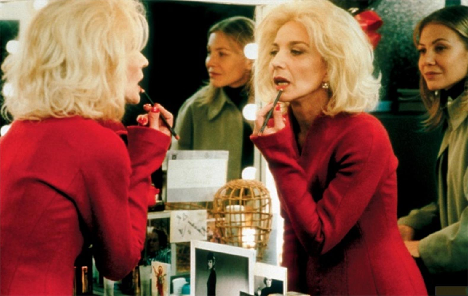 Una donna bionda si applica il rossetto davanti a uno specchio, con un'altra donna che guarda.