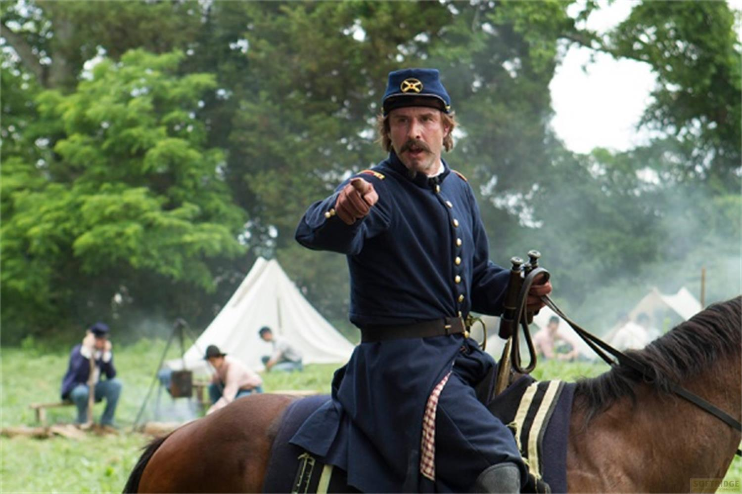 Mann in blauer Uniform auf Pferd zeigt nach vorne. Zelte im Hintergrund.