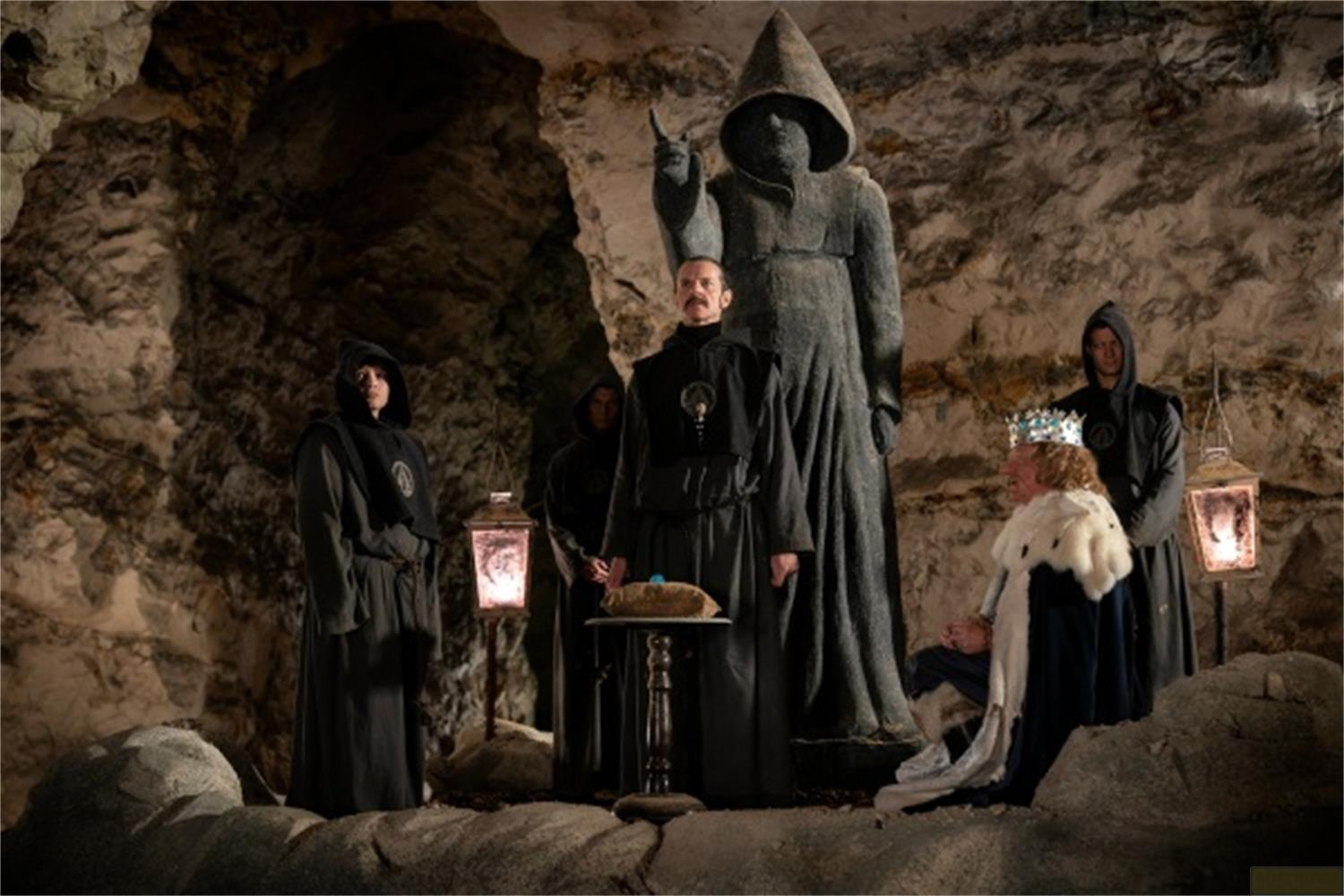 Menschen in Kapuzenroben in einer Höhle vor einer Steinstatue und einer Königin.