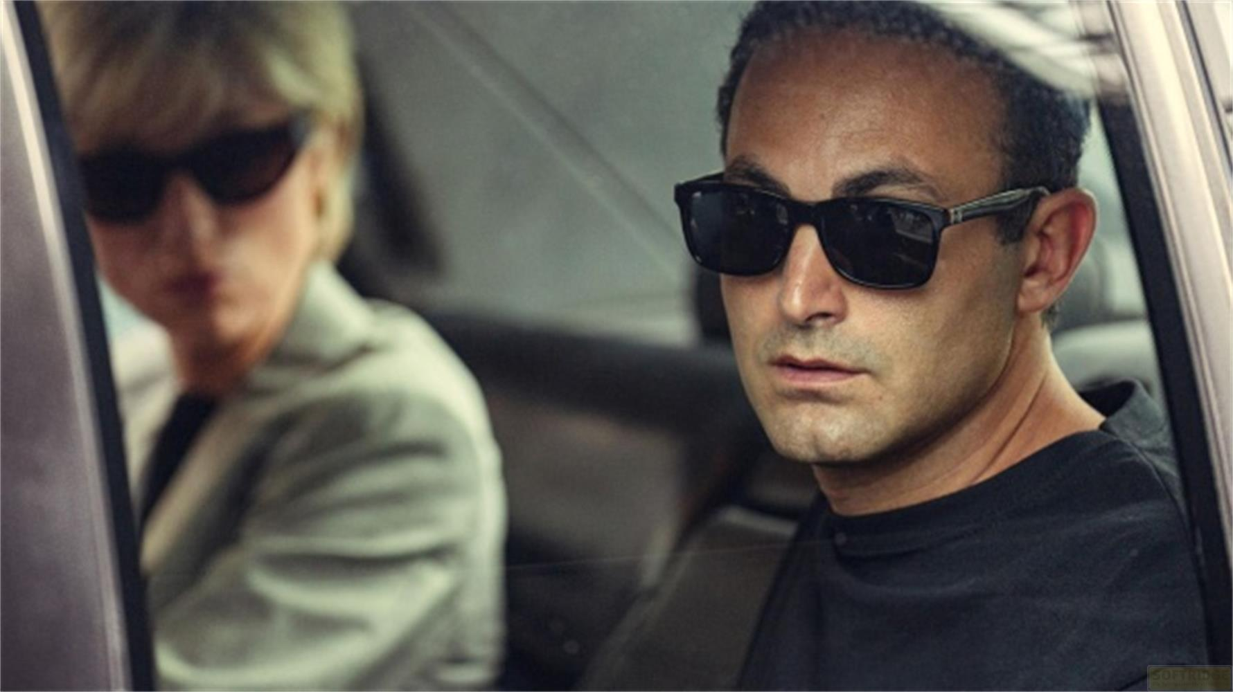 Uomo con occhiali da sole in primo piano, donna sfocata sullo sfondo in auto.