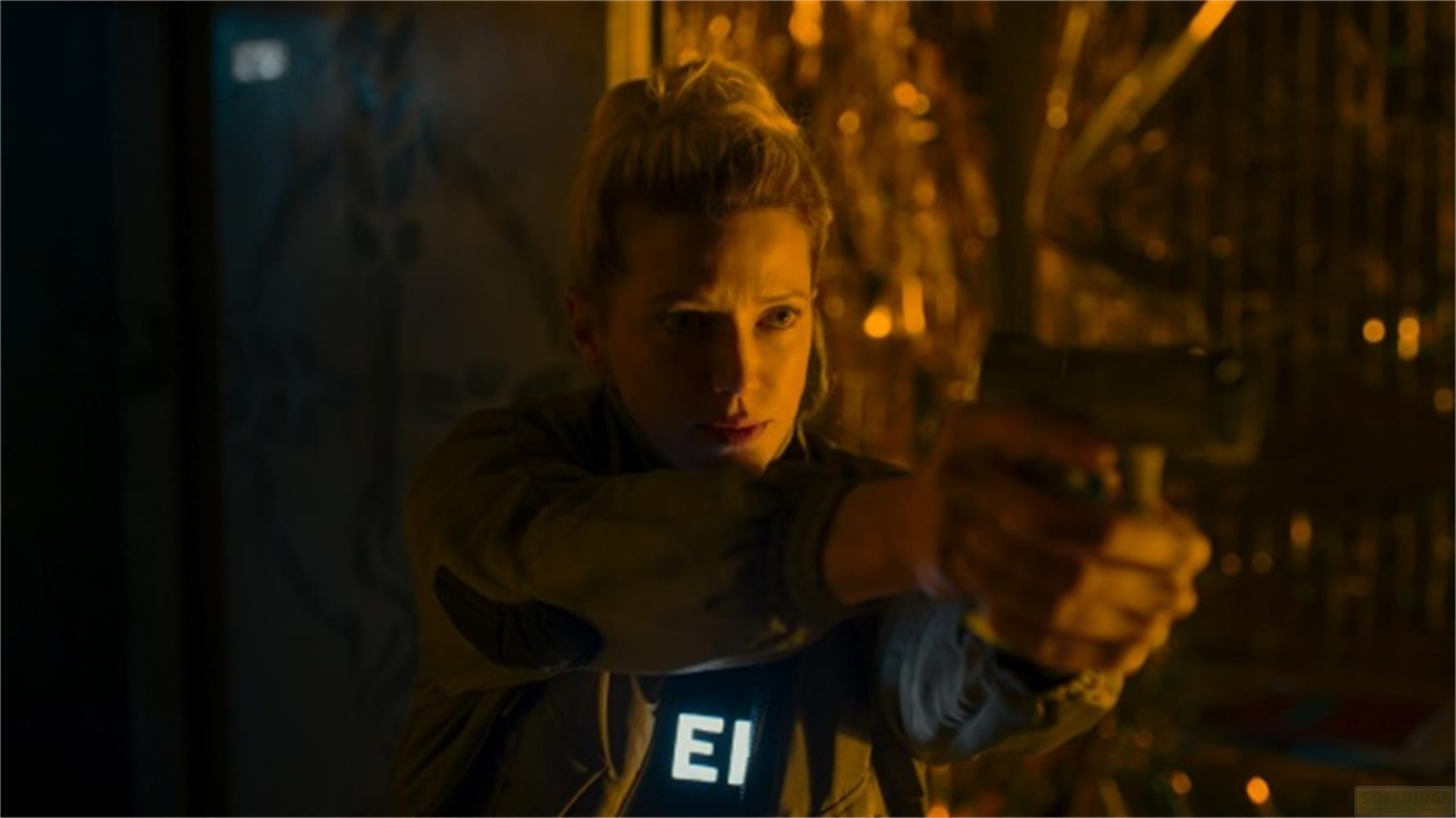 Una donna bionda punta una pistola, con "EI" che brilla sul petto.