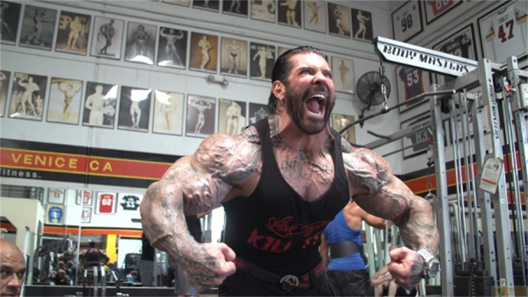 Un uomo muscoloso con tatuaggi urla in una palestra. Foto vintage di fitness adornano le pareti.
