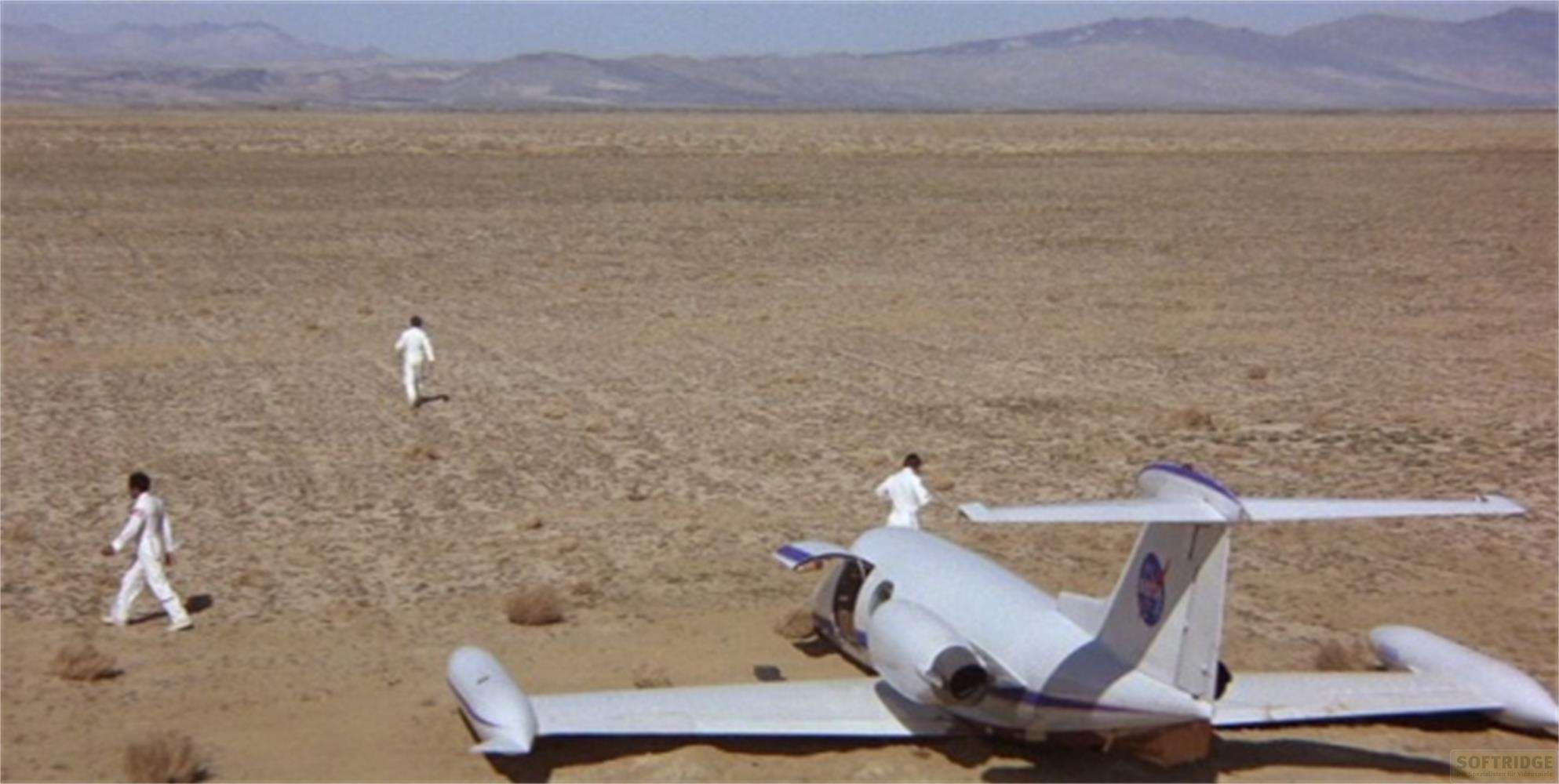 Tre persone in tuta bianca camminano su un paesaggio desertico verso un aereo caduto.