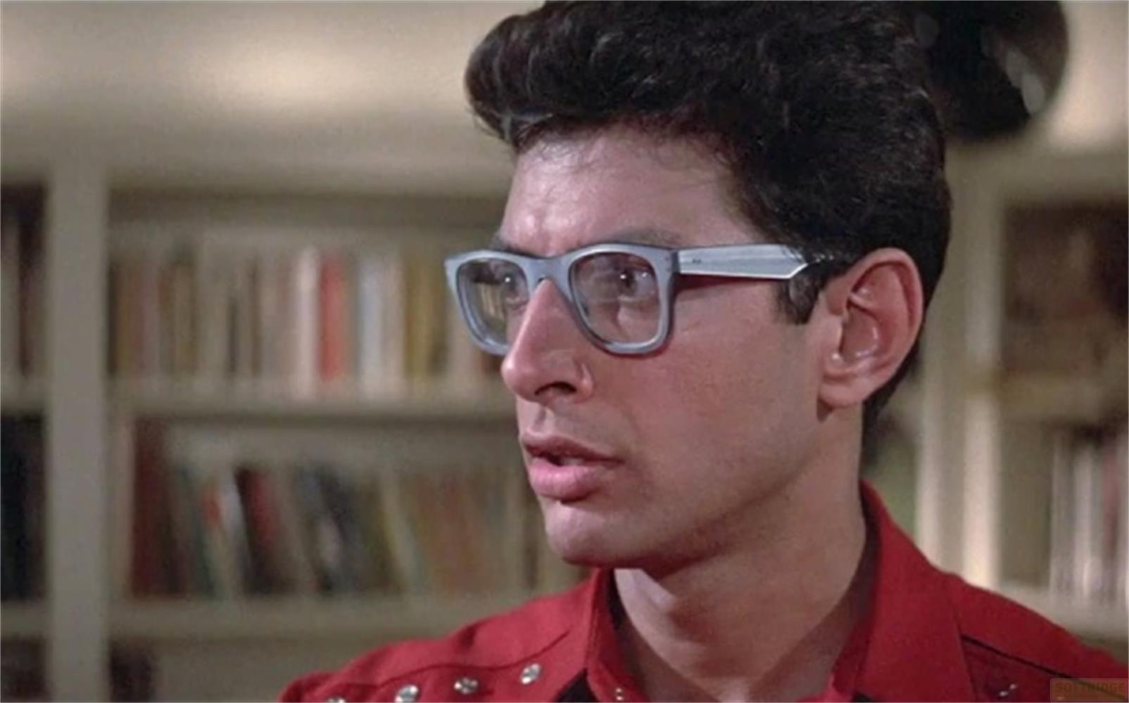 Un homme portant des lunettes et une chemise rouge devant une étagère.