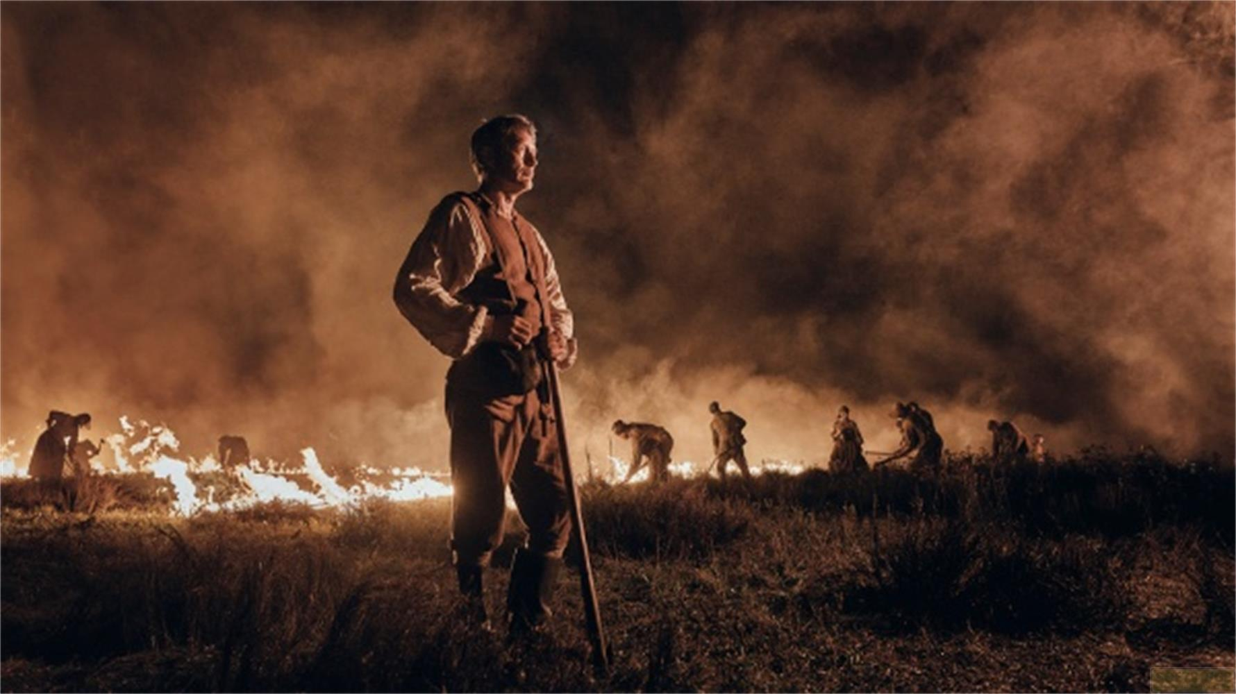 Un uomo con bastone davanti a un campo in fiamme, altri lavorano.