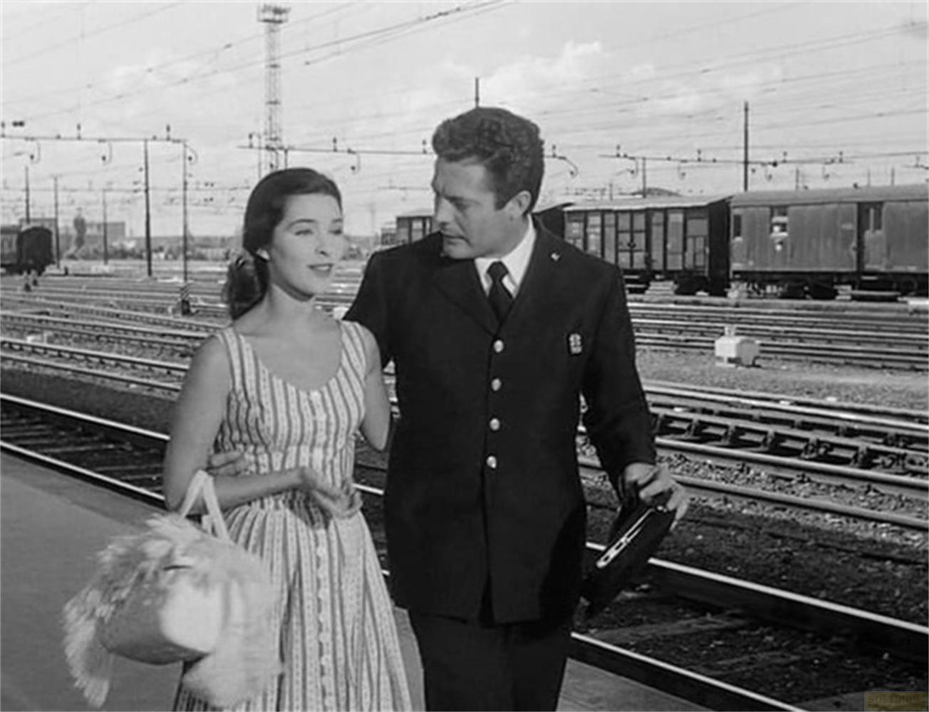 Uomo in divisa e donna con borsa camminano su binario, treni sullo sfondo.