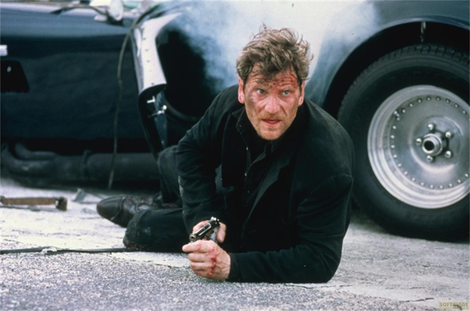Un uomo in nero si accovaccia accanto a un'auto con una pistola.