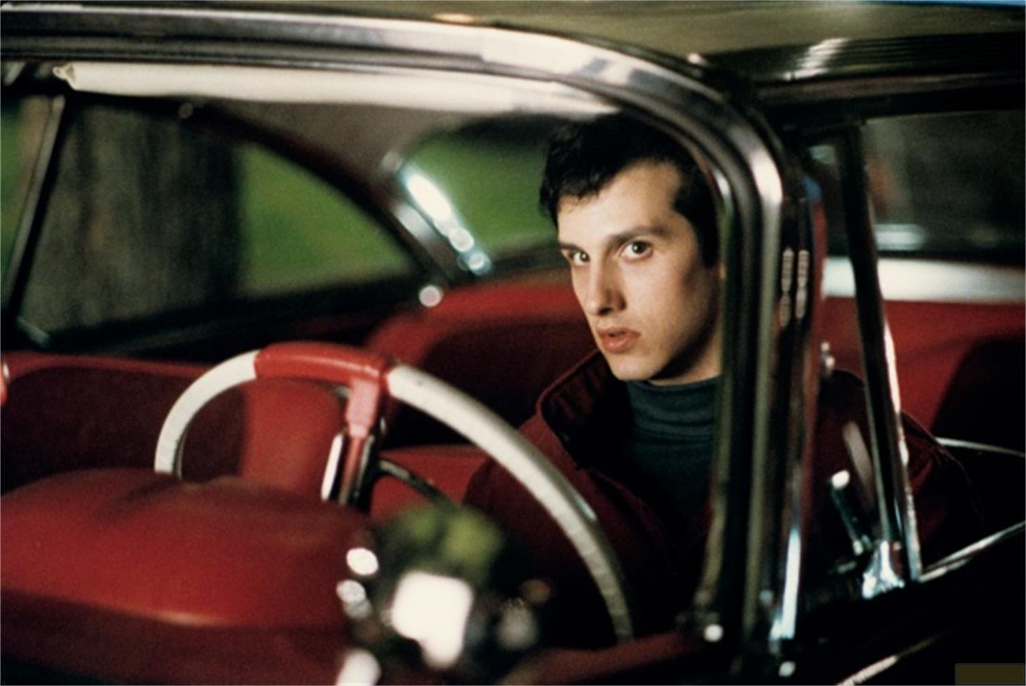 Un jeune homme regarde à droite depuis l'intérieur d'une voiture ancienne rouge.