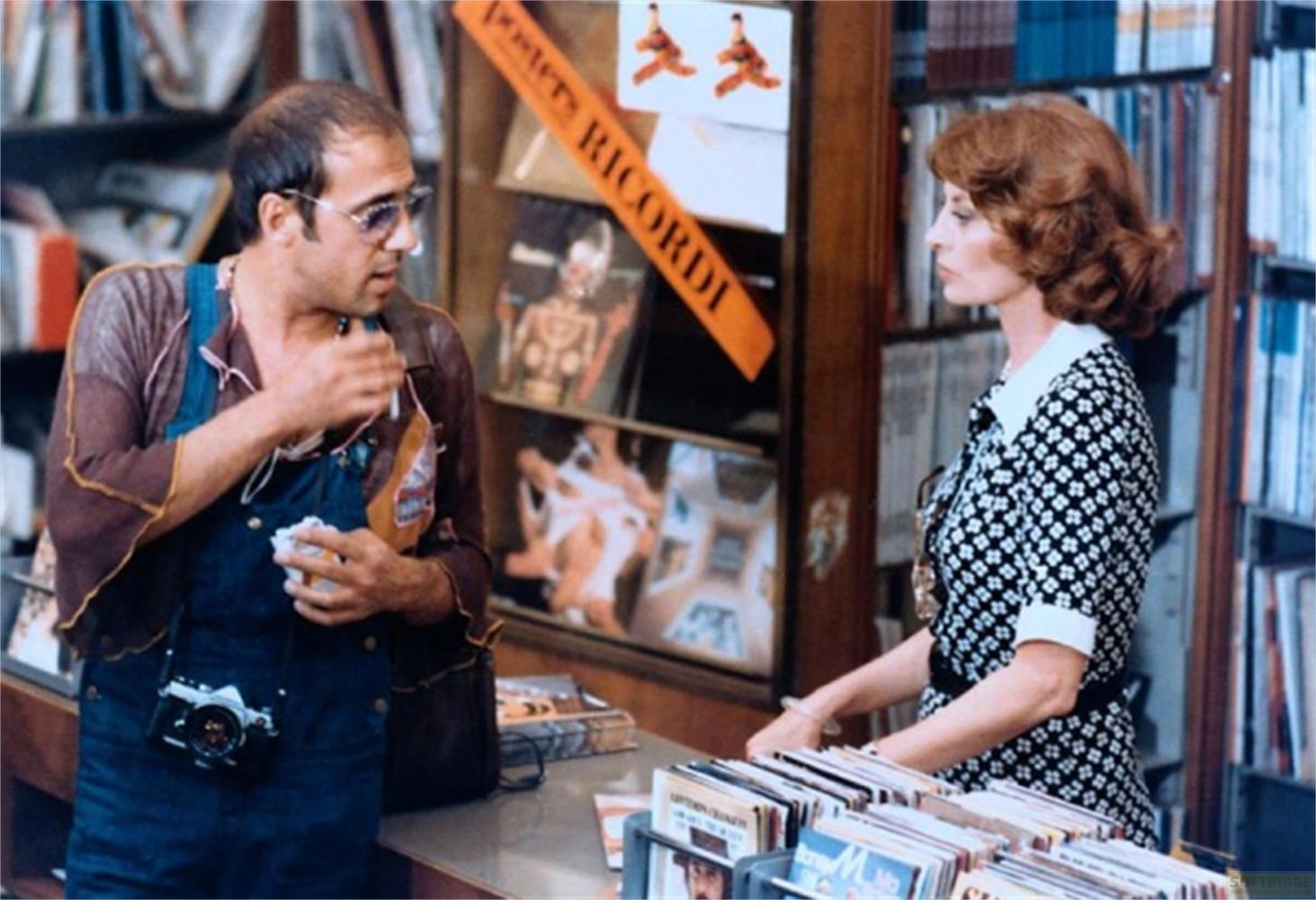 Uomo con occhiali e tuta parla con donna in abito a fantasia davanti a librerie.
