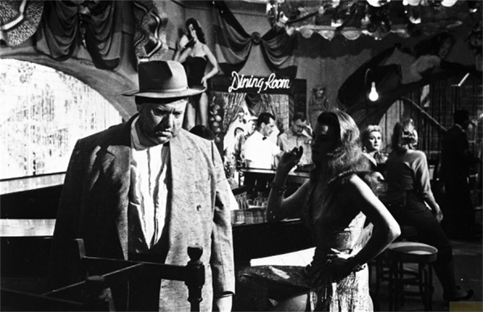 Un homme au chapeau est au premier plan. Une femme aux longs cheveux est assise au bar.