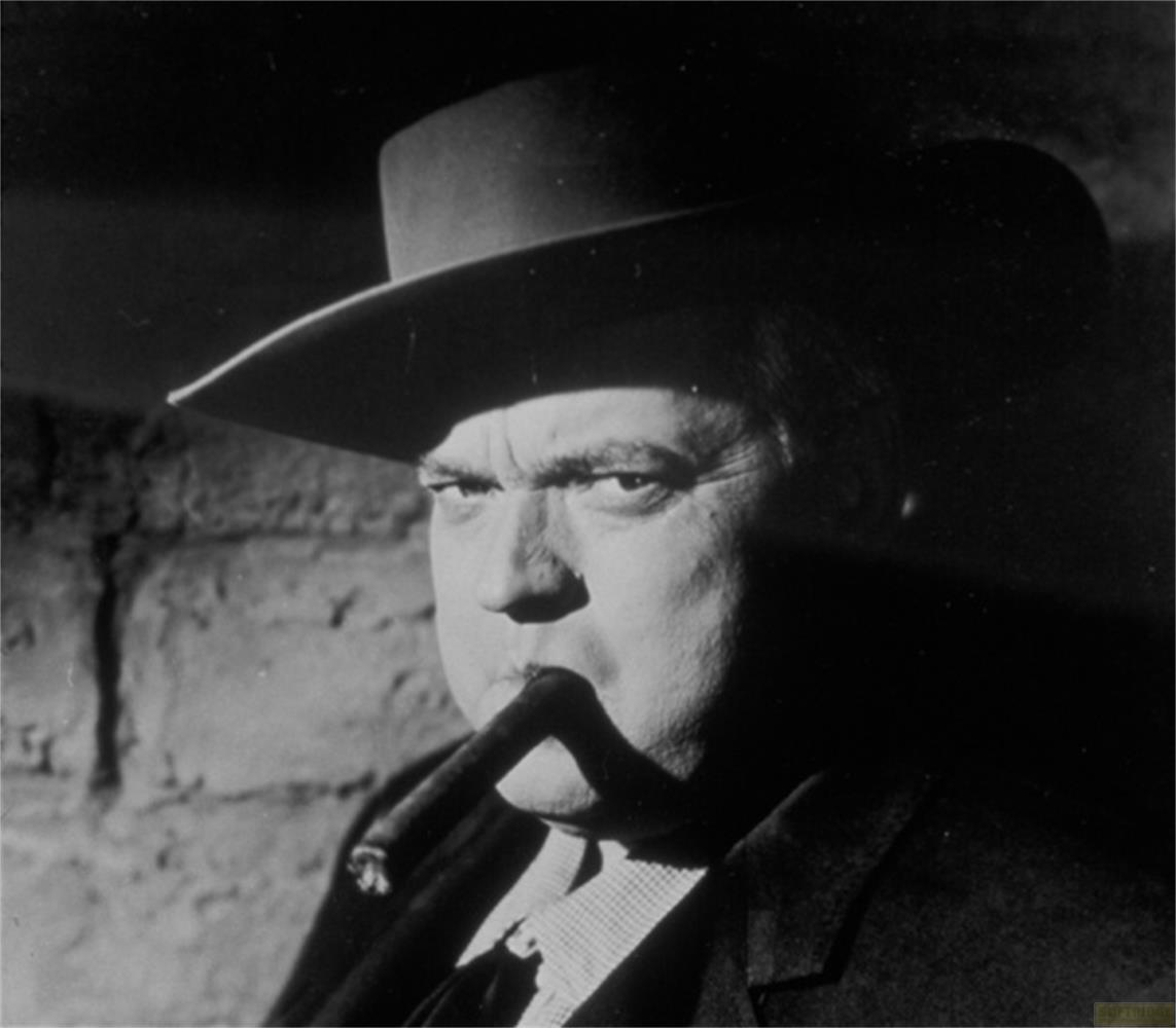 Un homme portant un chapeau fume un cigare, avec un mur de briques en arrière-plan.