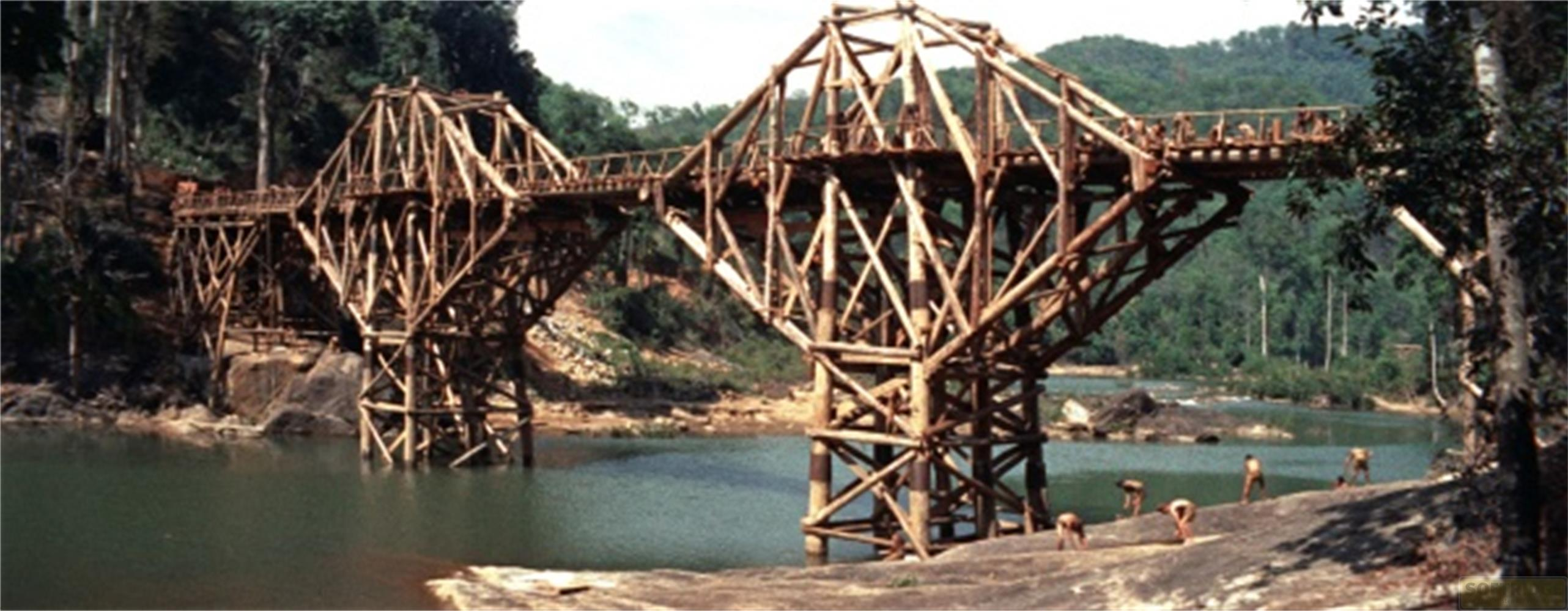 Un ponte a traliccio in legno in costruzione su un fiume, con operai sulla riva.