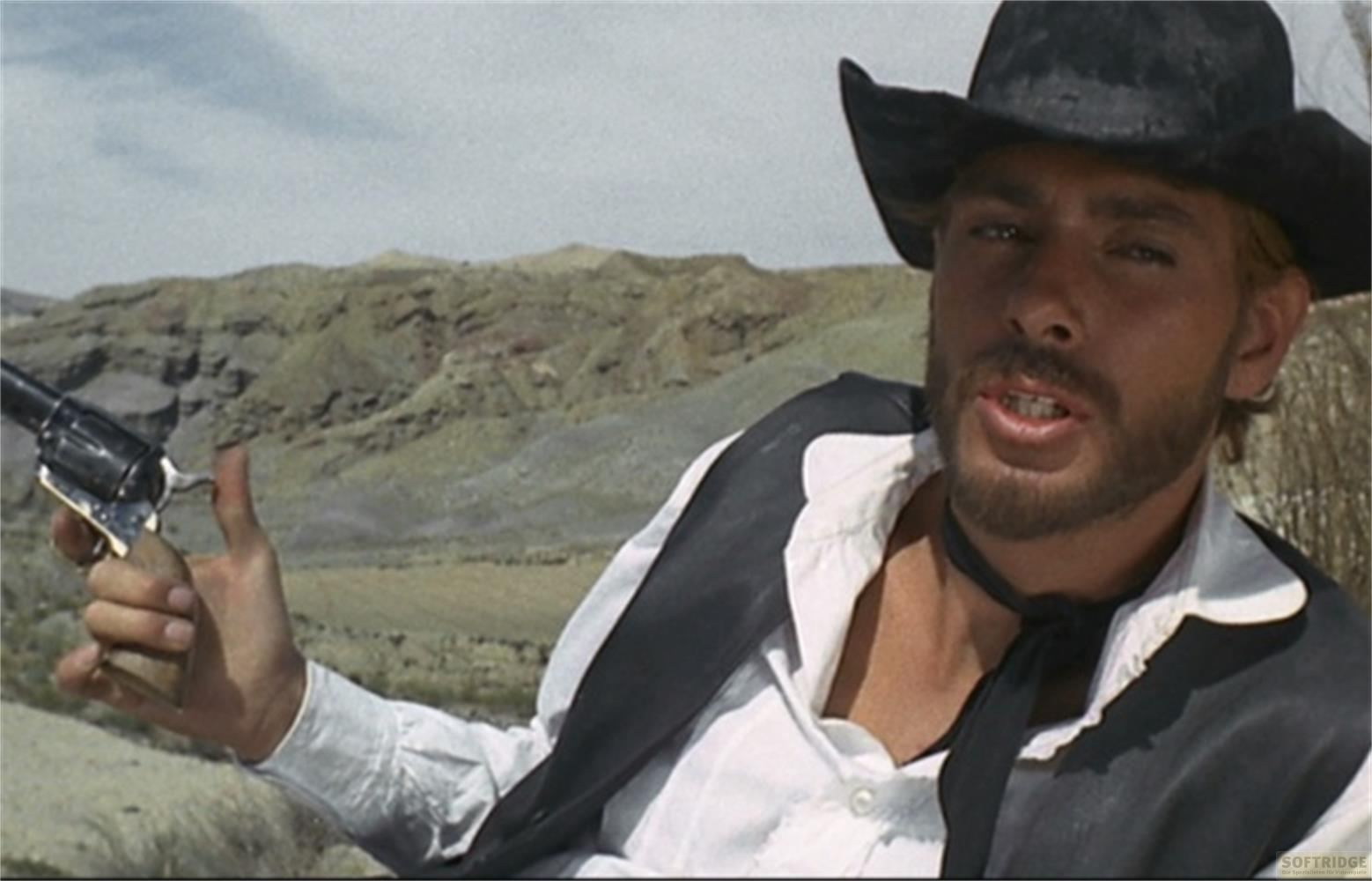 Mann mit Hut hält Revolver in felsiger, trockener Landschaft.