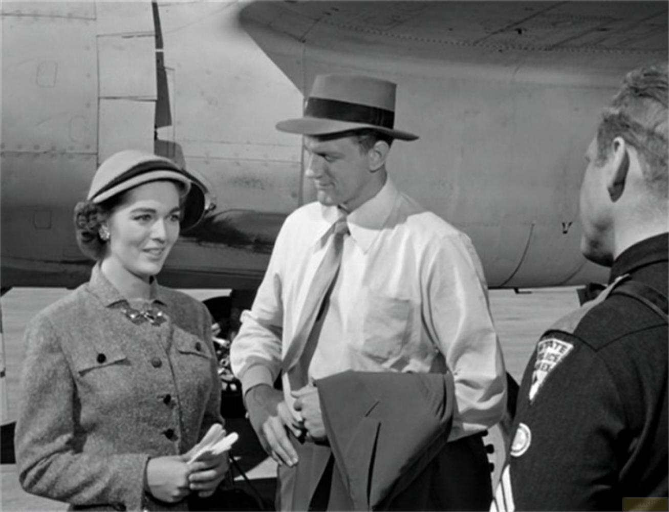 Una donna con cappello e tailleur parla con un uomo con cappello e abito vicino a un aereo.