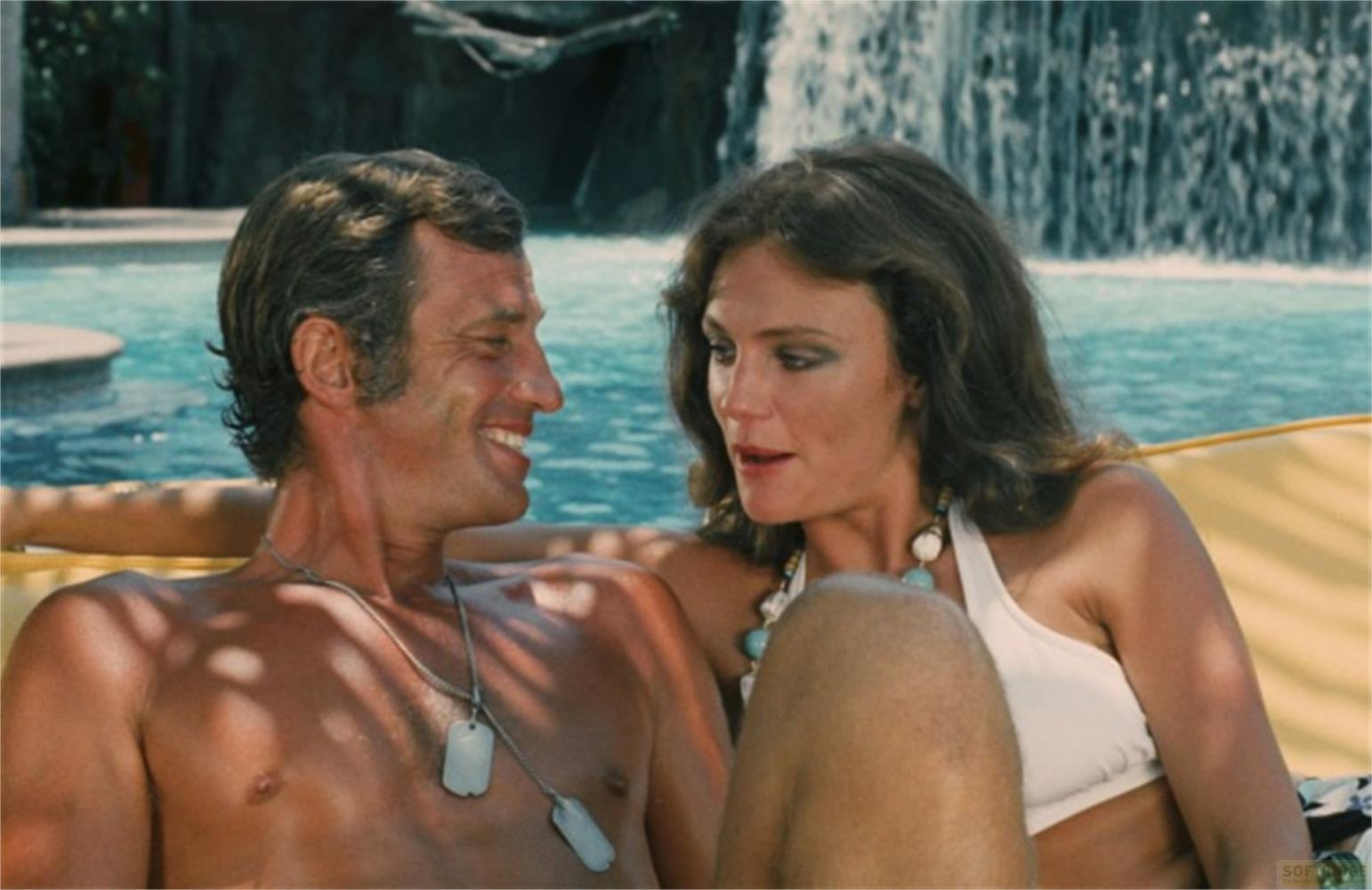 Ein Mann und eine Frau sitzen am Pool mit einem Wasserfall im Hintergrund.