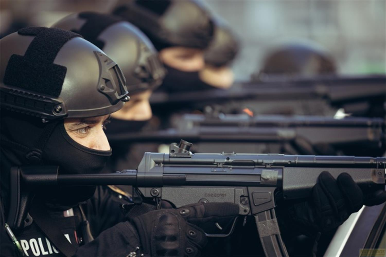 Uomo in tenuta nera e casco che impugna un'arma da fuoco. Diverse persone dietro.