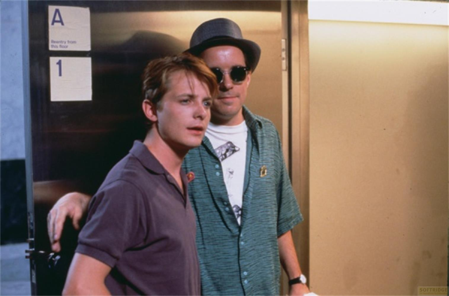 Deux hommes devant une porte. L'un porte un polo violet, l'autre un chapeau, des lunettes de soleil et une chemise.