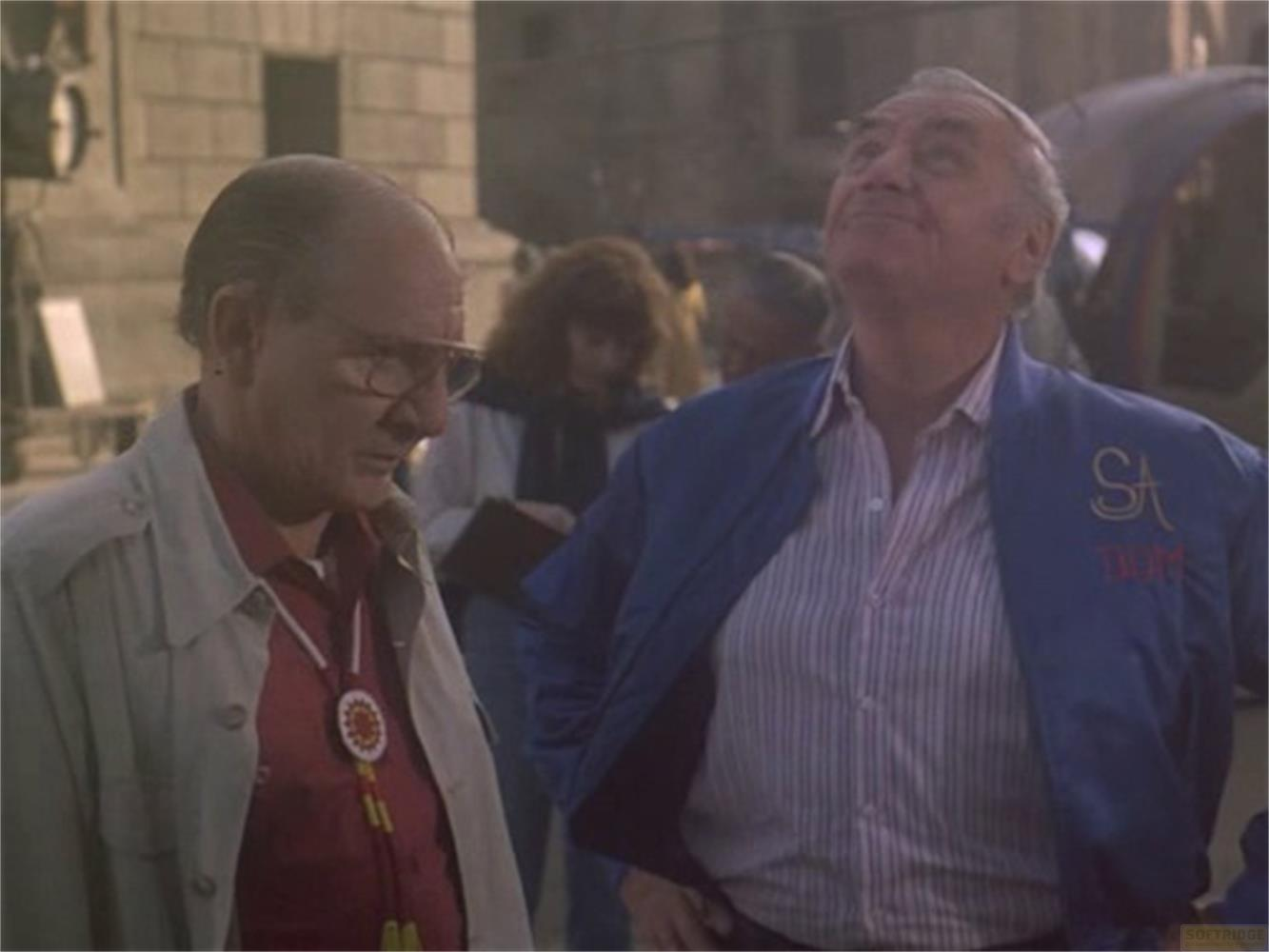 Due uomini all'aperto, uno con camicia rossa e collana, l'altro con giacca blu.