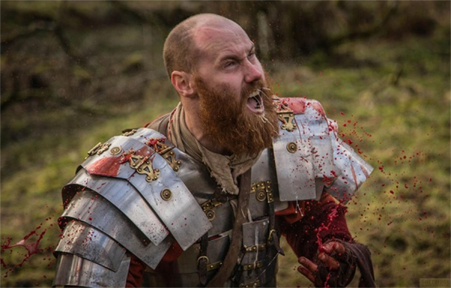 Un uomo in armatura con barba sputa sangue. Il sangue schizza sull'armatura.