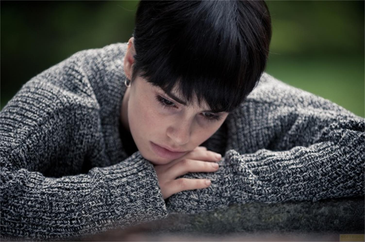 Una persona giovane con capelli neri corti, maglione grigio, appoggia la testa sulle braccia.