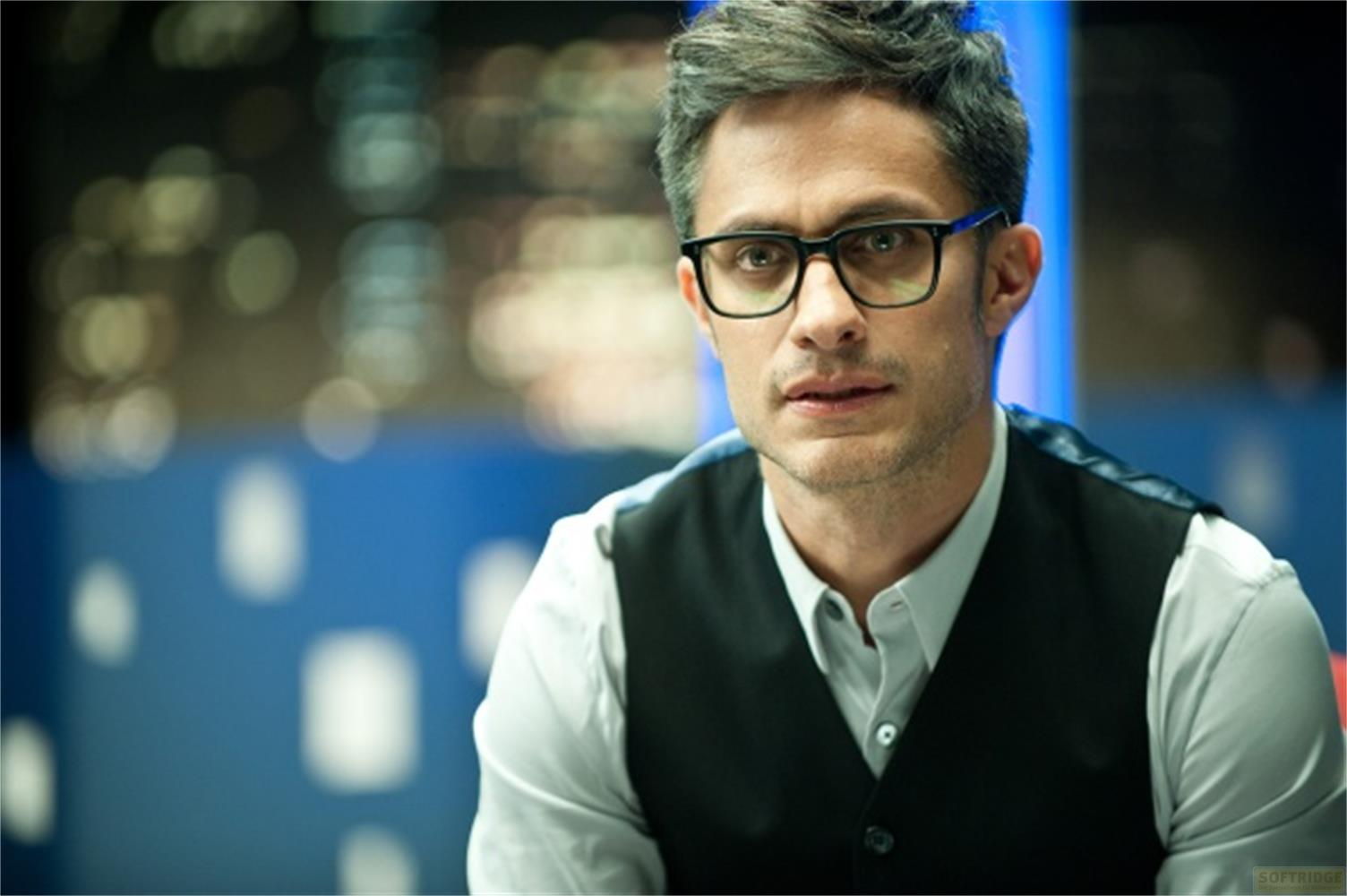 Uomo con occhiali e gilet su camicia, sfondo blu sfocato.