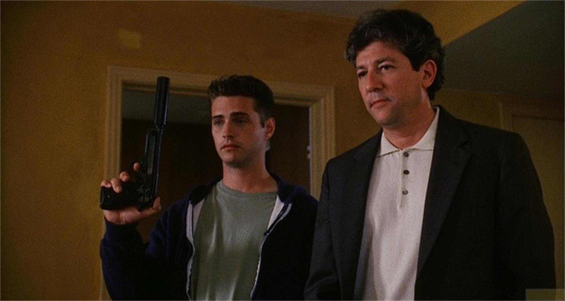 Un uomo con capelli scuri tiene una pistola. Accanto a lui c'è un uomo in giacca.