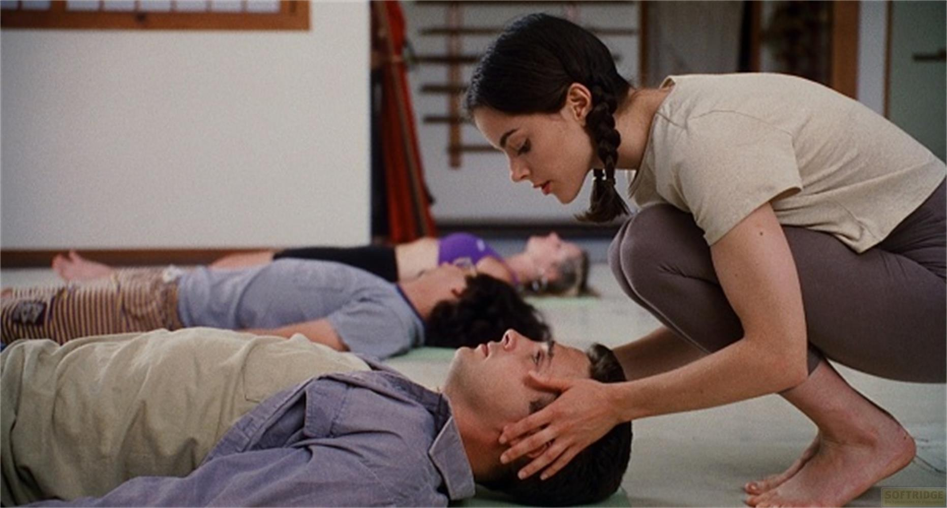 Una donna in abiti da yoga si inginocchia su un uomo sdraiato, toccandogli delicatamente la testa.