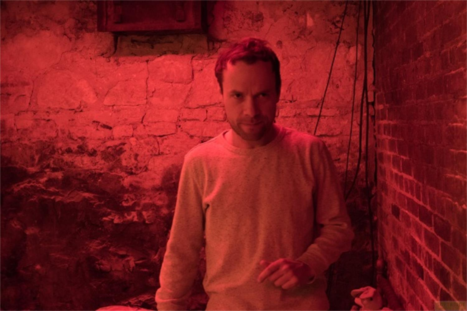 Ein Mann in einem grauen Pullover steht vor einer Ziegelwand, beleuchtet von rotem Licht.