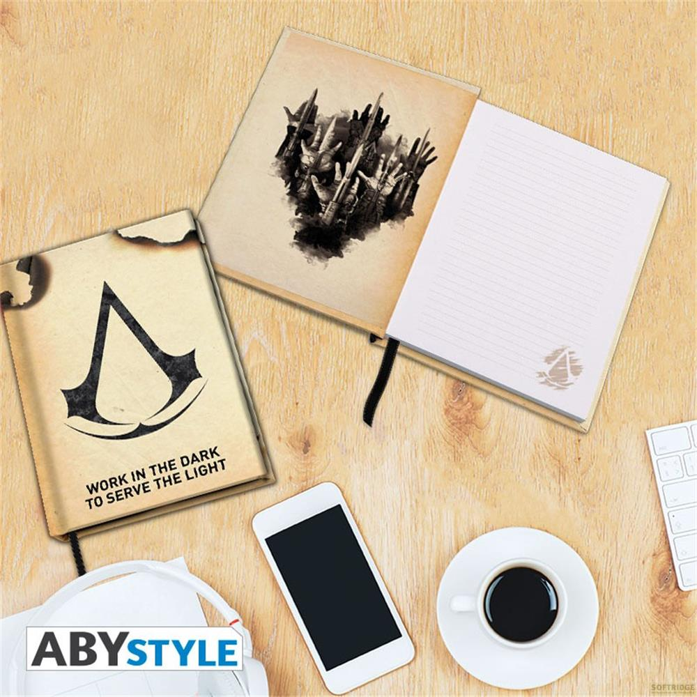 Ein Assassin's Creed Notizbuch mit Logo, ein geöffnetes Notizbuch mit Händen, ein Handy, Kopfhörer und Kaffee.
