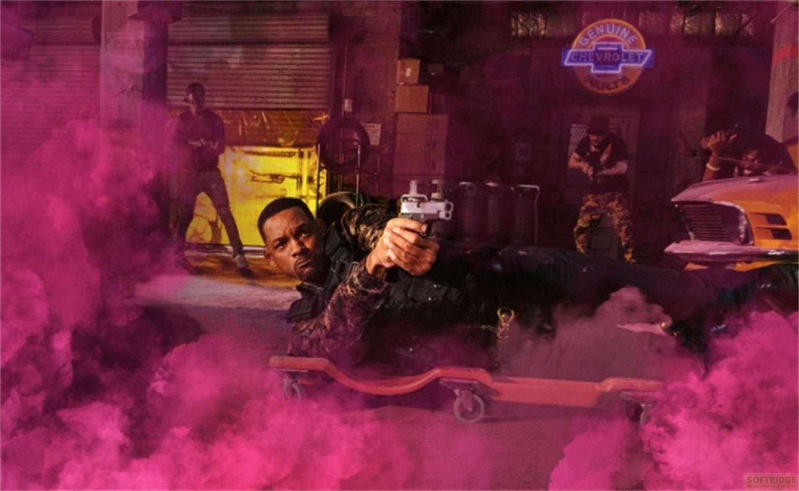 Will Smith giace su un carrello, sparando con una pistola, circondato da fumo rosa.