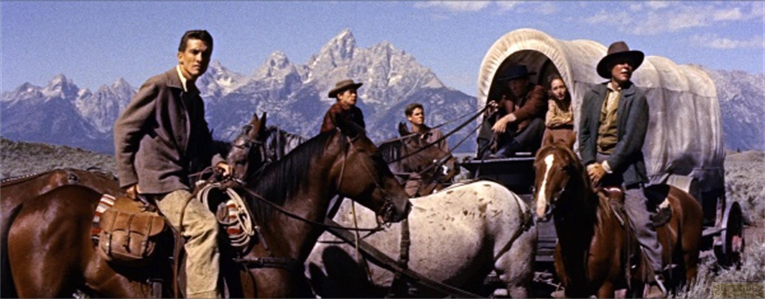Cavalli e carro coperto davanti alle montagne, uomini a cavallo, un uomo seduto sul carro.