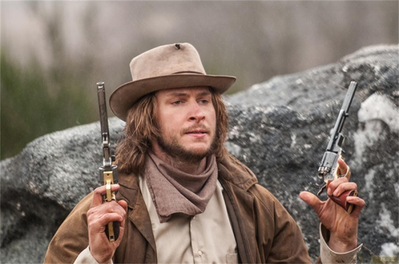 Un homme en tenue western, chapeau et armes, près des rochers.