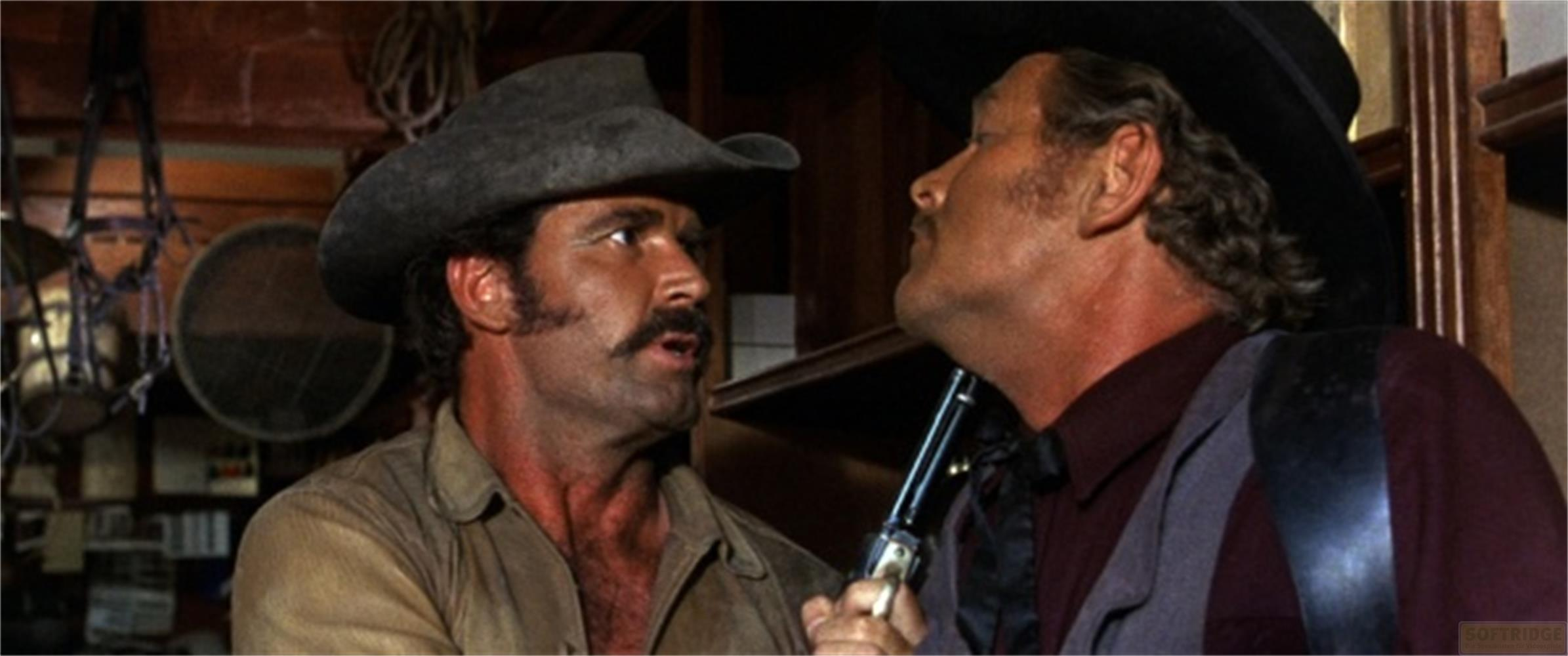 Due uomini in abiti western con cappelli in primo piano, una briglia e scaffali sullo sfondo.