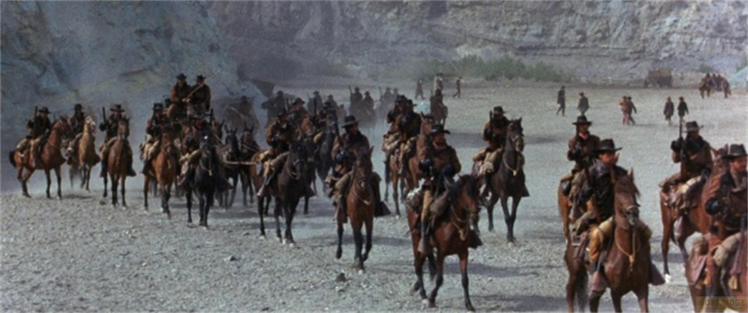 Un gruppo di cowboy cavalca attraverso un paesaggio polveroso e roccioso.