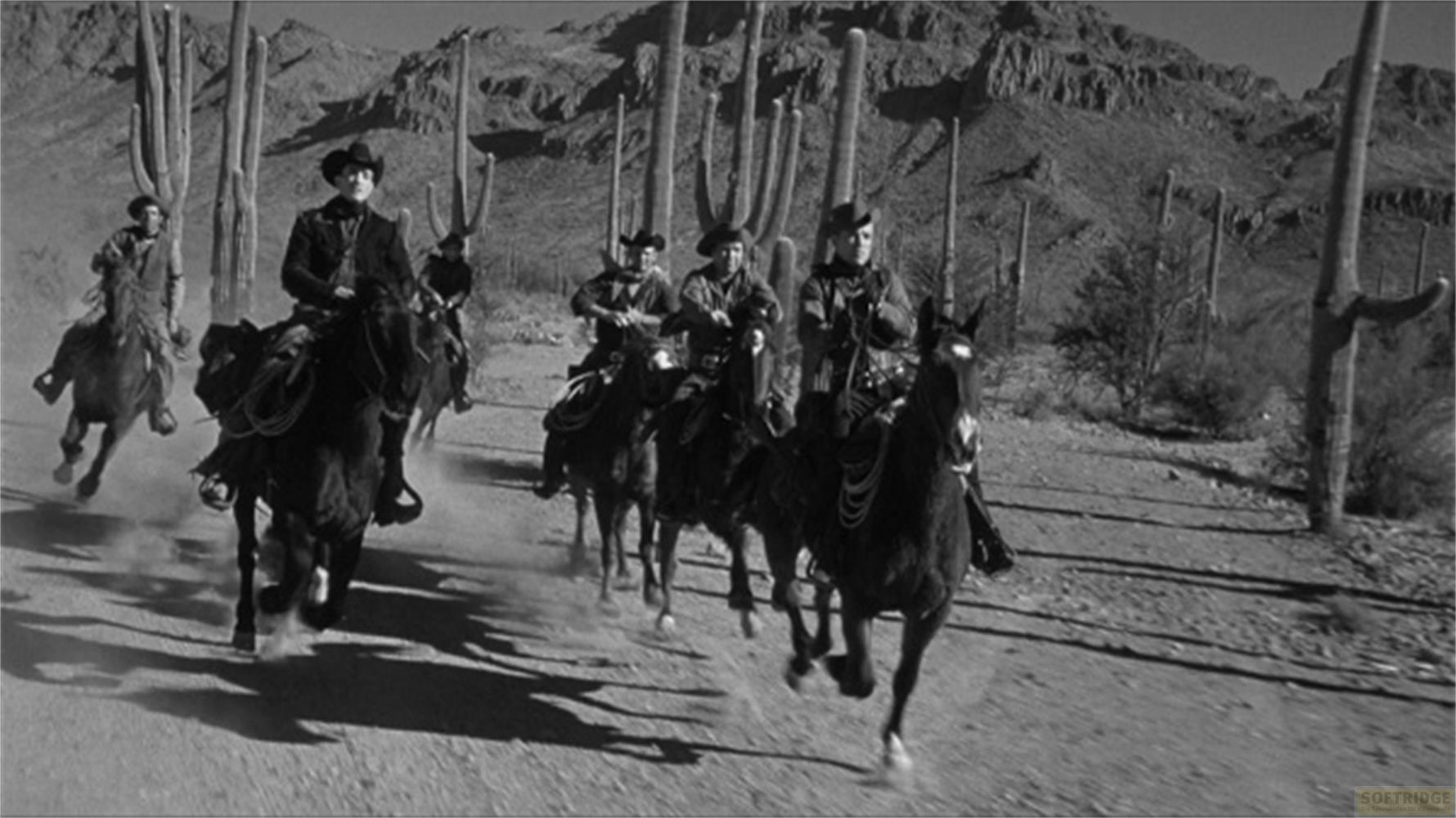 Männer in Cowboy-Kleidung reiten auf Pferden durch eine Wüstenlandschaft mit Kakteen.