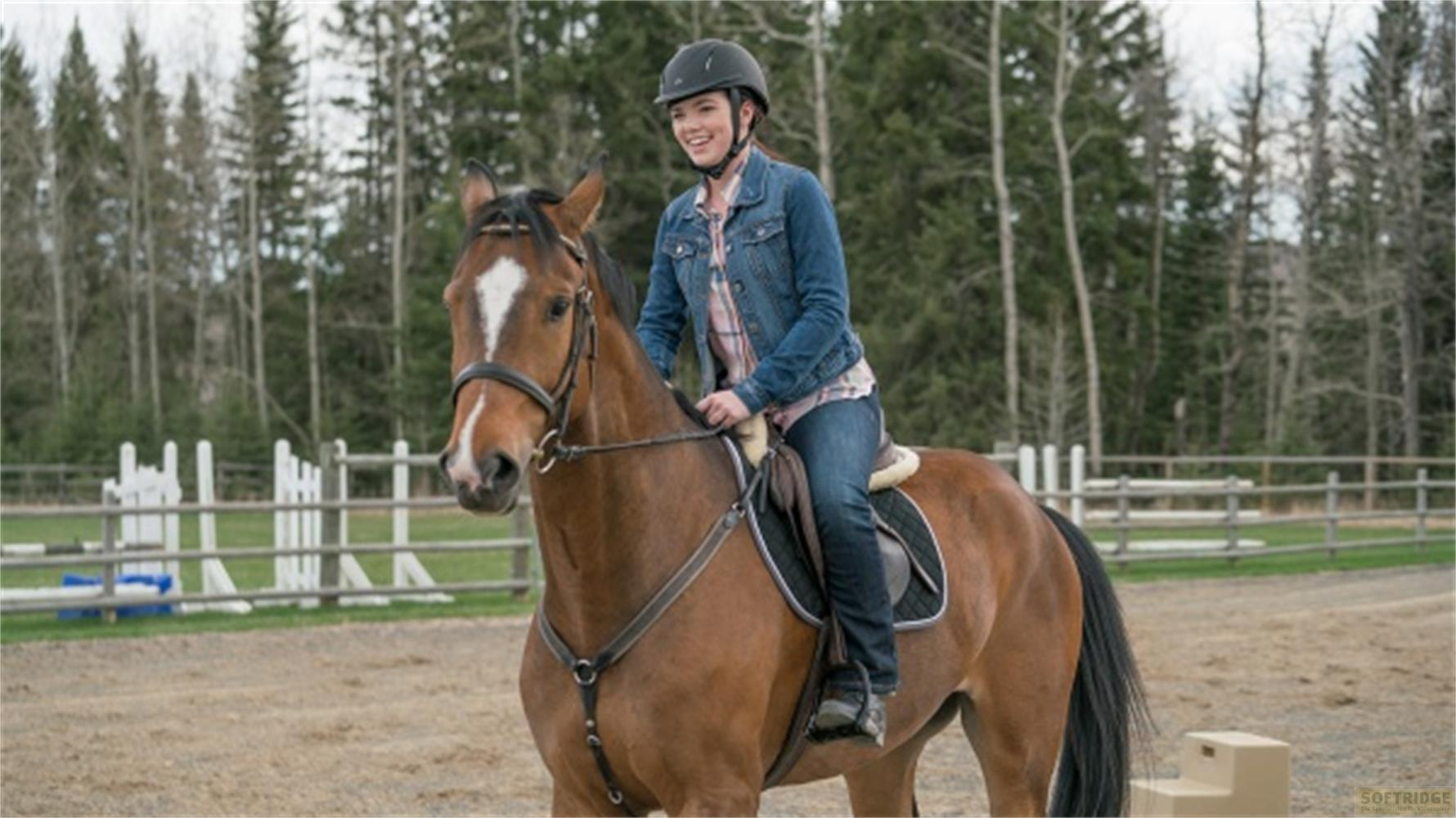 Una giovane donna sorridente con casco cavalca un cavallo marrone in un'arena recintata con alberi sullo sfondo.