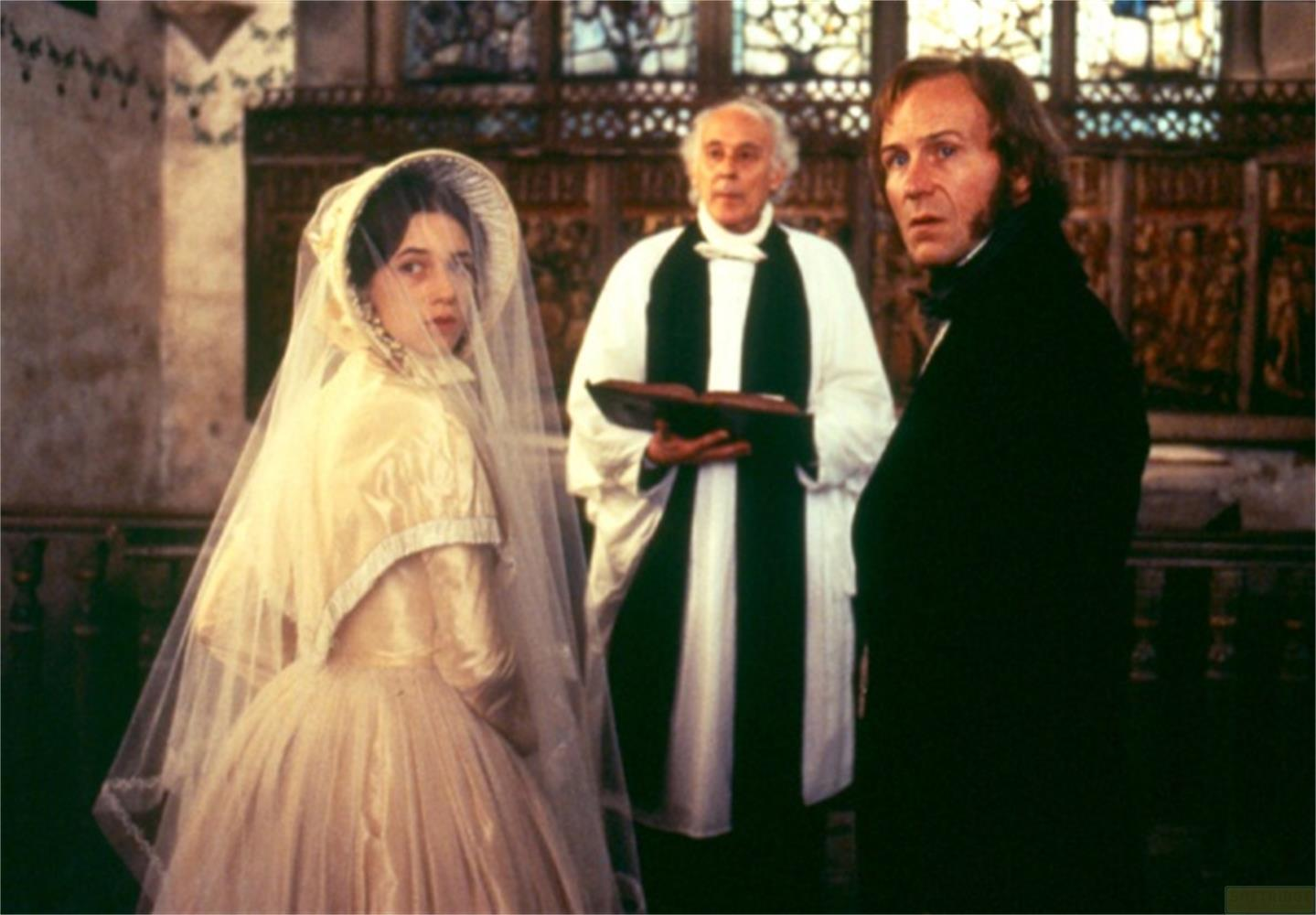 Una sposa in abito color crema con velo sta accanto a un ecclesiastico e un uomo in abito nero.