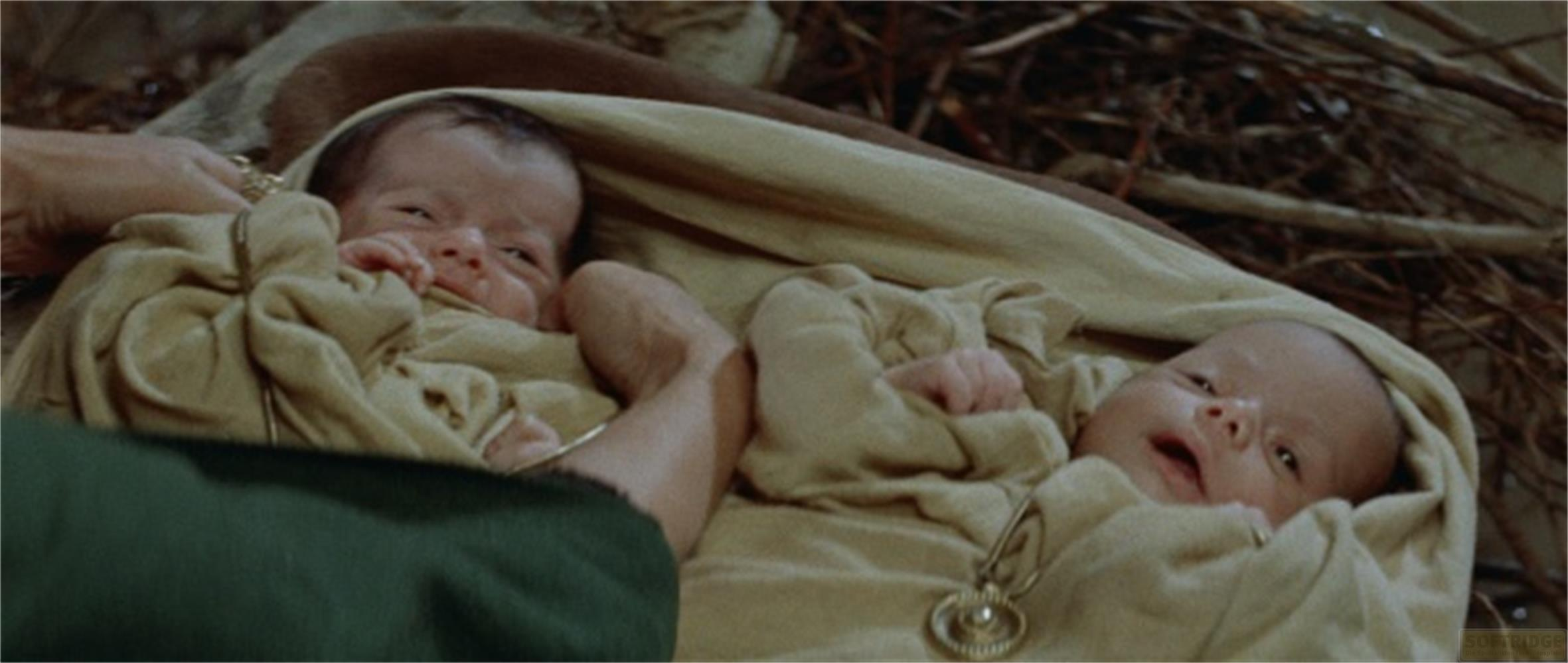 Due neonati avvolti in panni beige, uno guarda in alto, una mano vicina.