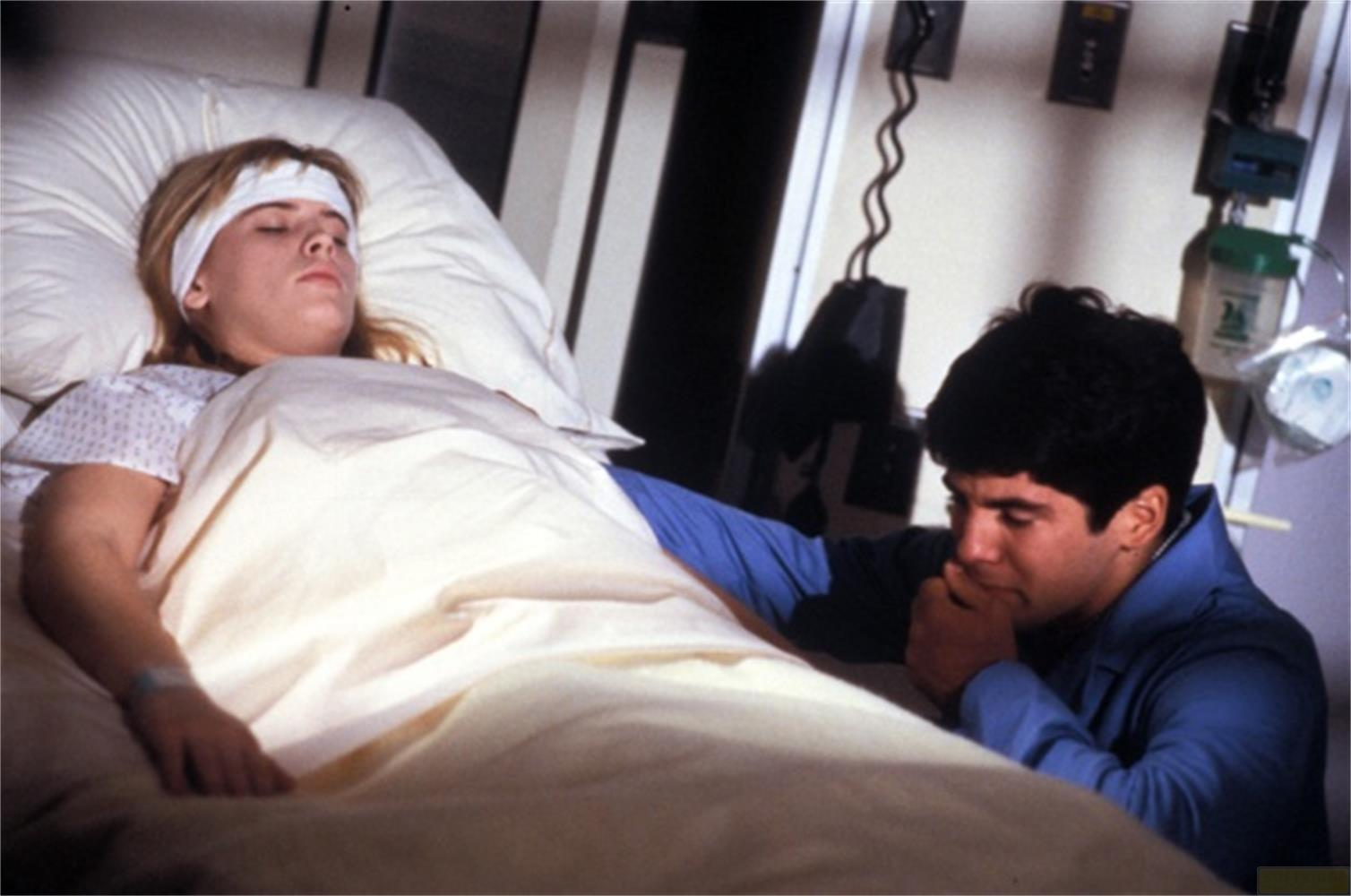 Uomo tiene la mano di una donna in letto d'ospedale con benda sulla testa.