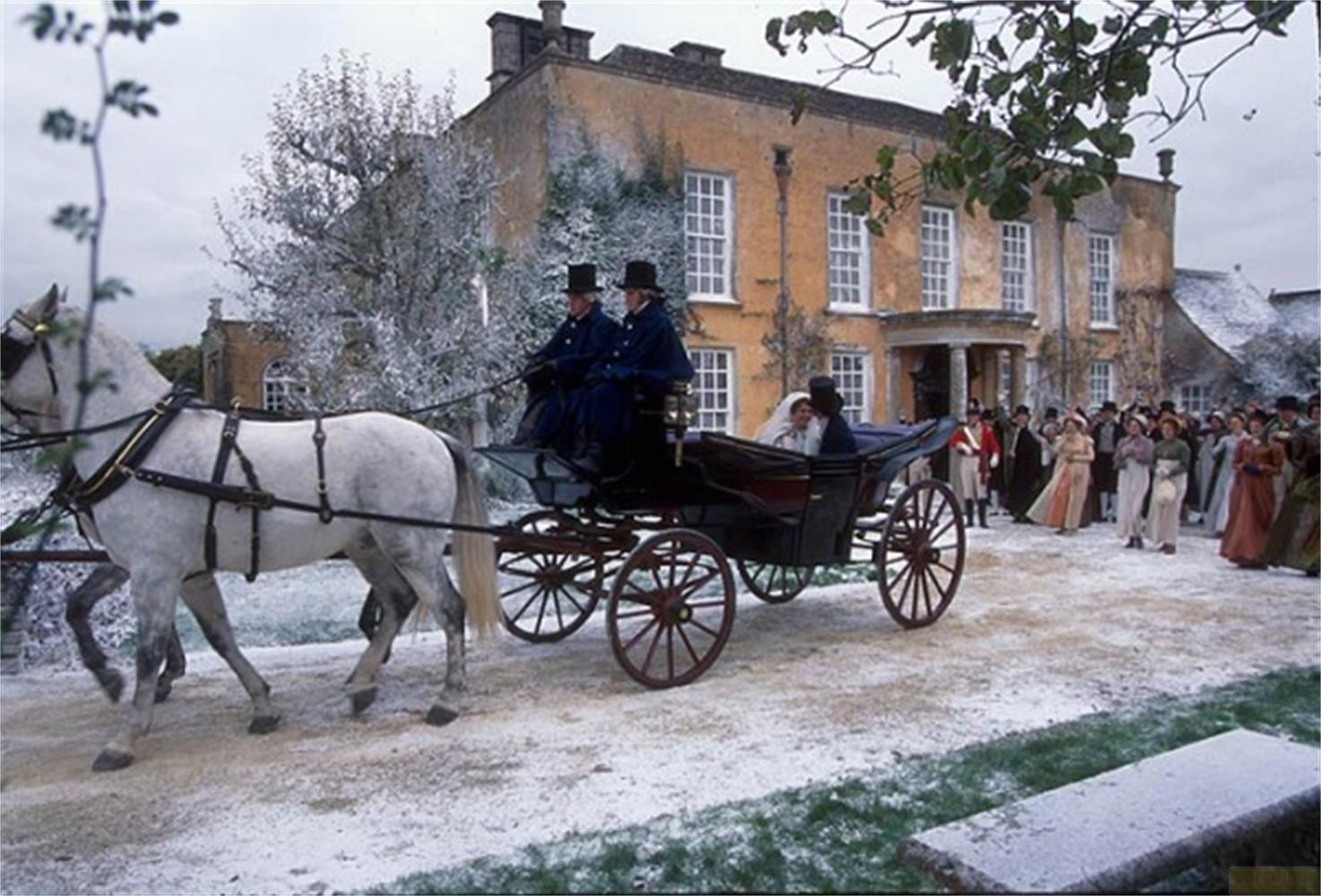 Une calèche tirée par des chevaux se déplace sur un chemin enneigé vers une maison, avec des gens en arrière-plan.